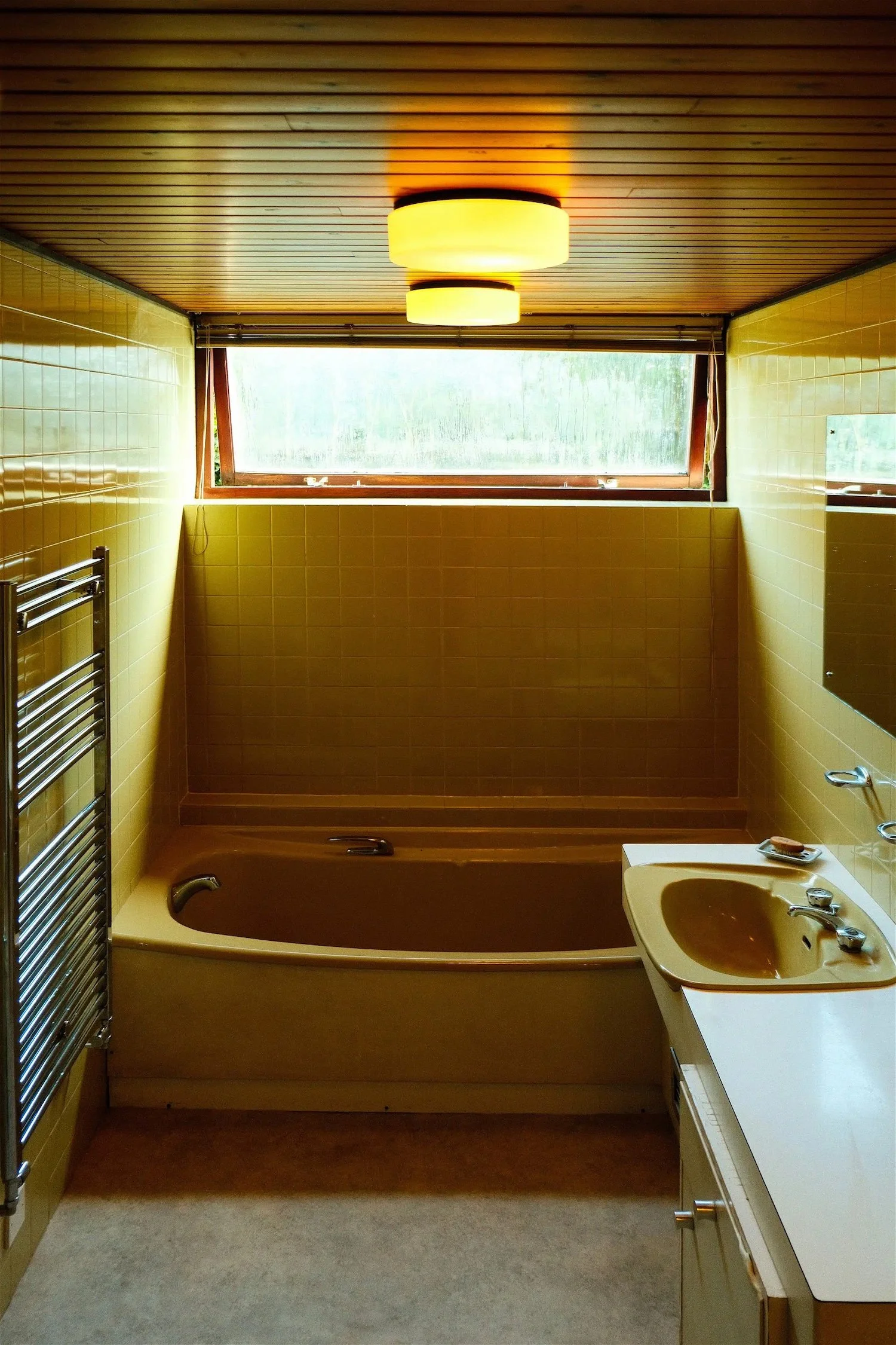 Vintage bathroom with yellow tiles, a built-in bathtub, a yellow sink, a mirror, and a window with a view of greenery outside.