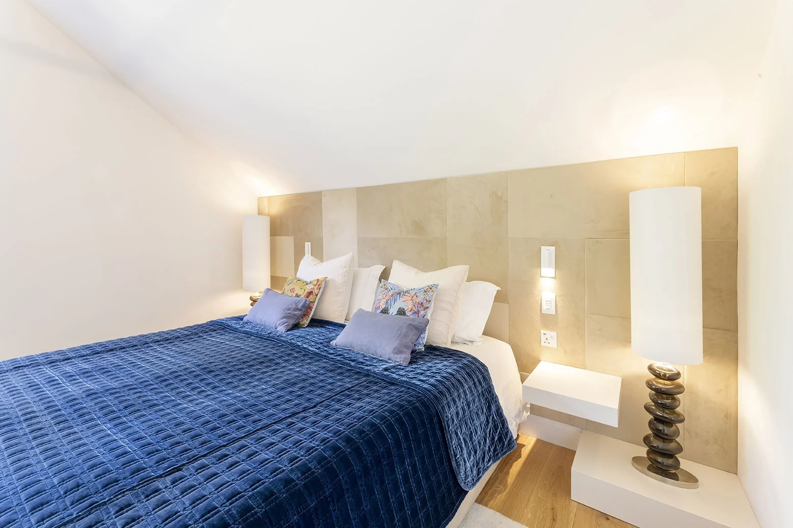 A bedroom with a large bed covered in a dark blue quilt, multiple pillows, and a wooden headboard. There are two tall lamps on either side of the bed and a white nightstand on the right side.