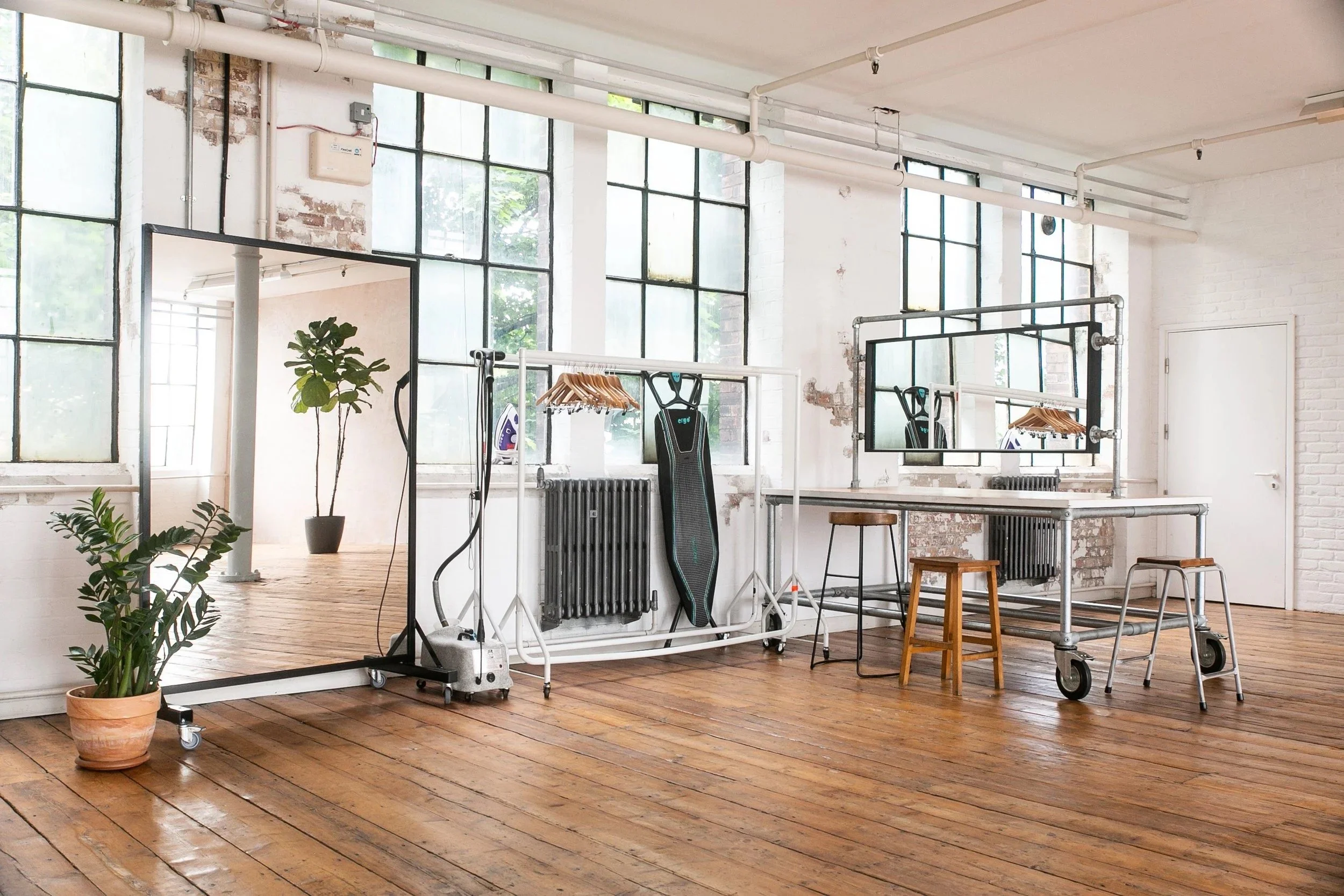 Empty creative workspace with large industrial windows, plants, mirrors, and furniture on wooden floors.