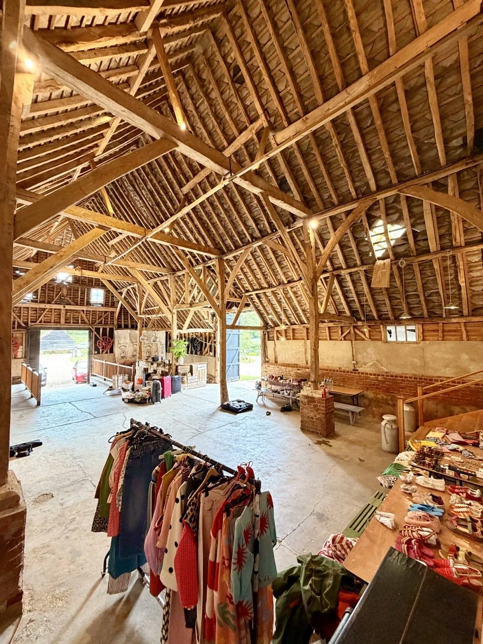 Interior of a rustic barn used for a flea market, with clothing on a rack in the foreground and tables with various items, including accessories and decorations, along one wall. Wooden beams and vaulted ceiling are visible.