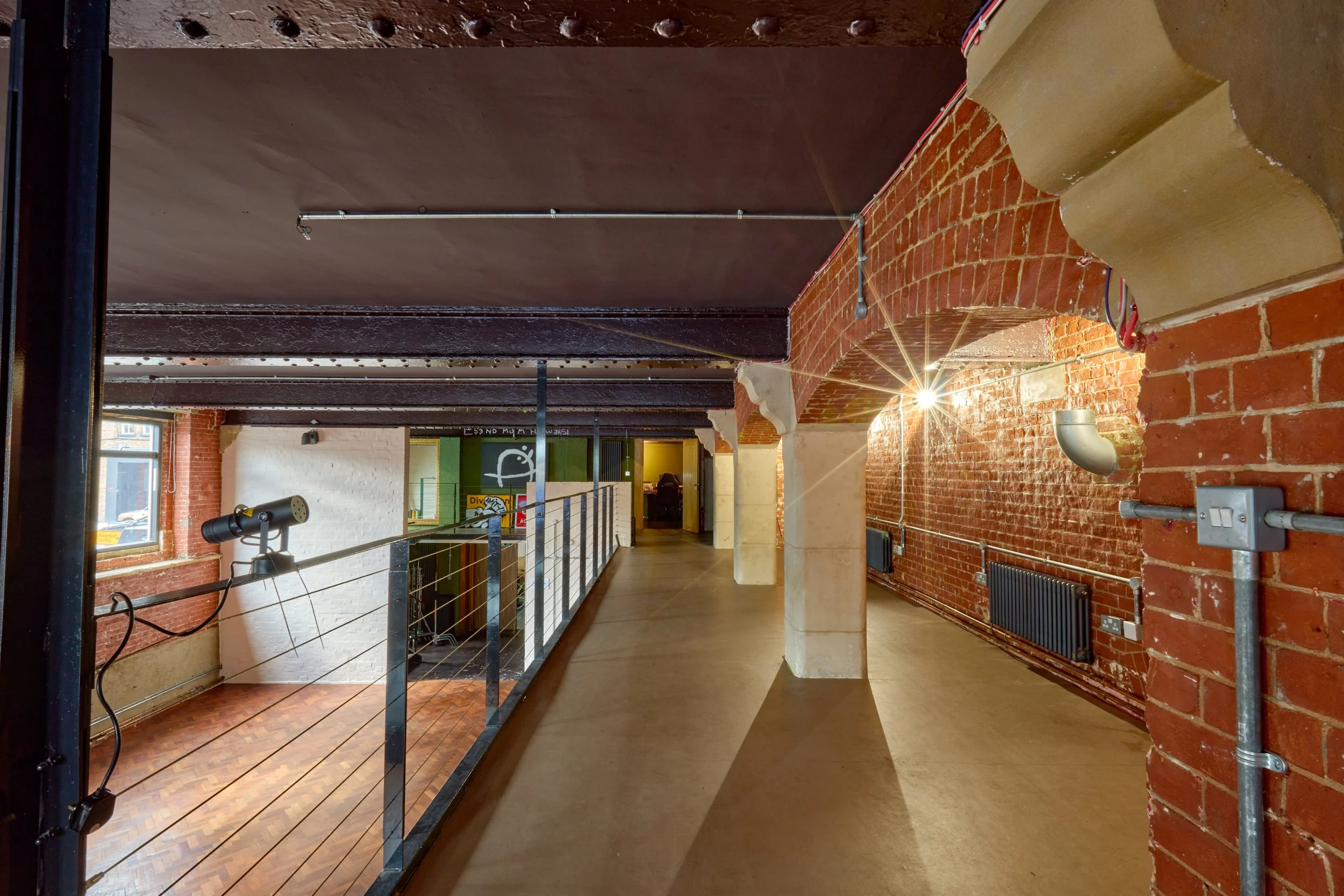 Interior of a building with exposed brick walls, arched brick ceiling, wooden flooring, and industrial lighting fixtures; a walkway with metal railings visible along the side.