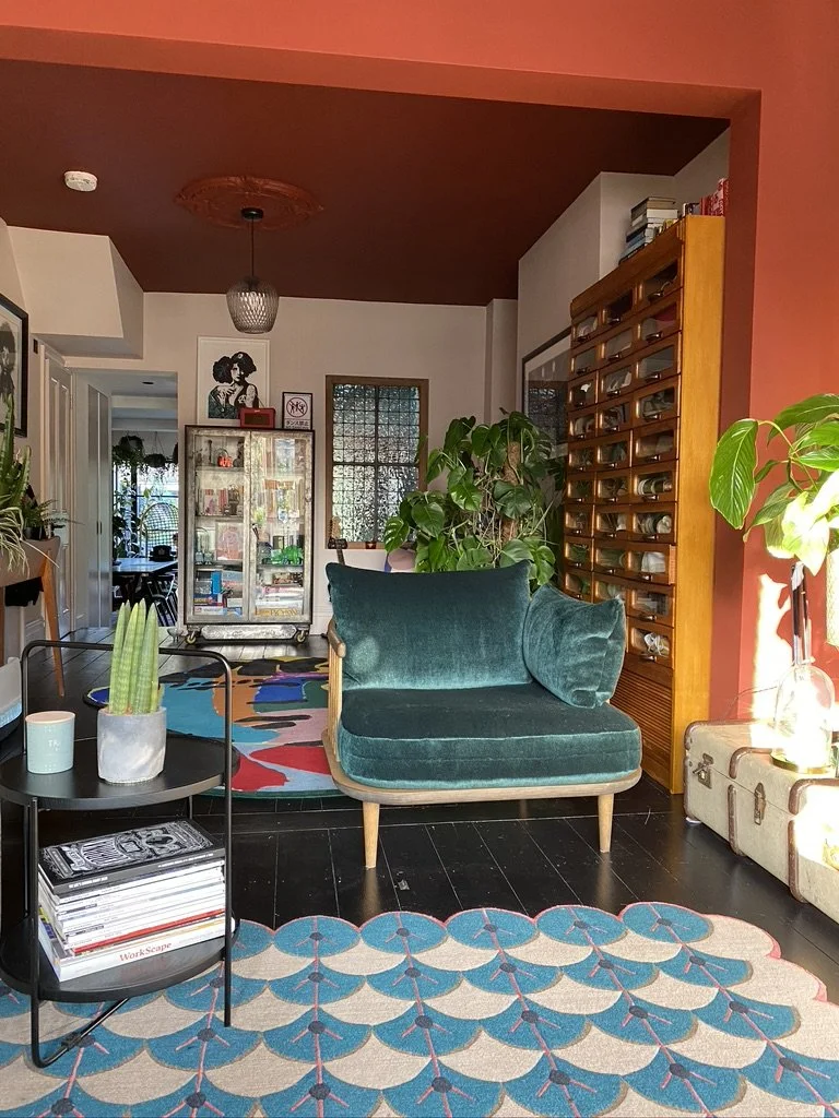 Living room with teal velvet loveseat, black side table with books and a potted cactus, colorful patterned rug, plants, and a cabinet of drawers, with artwork on the walls and a wicker ceiling light.