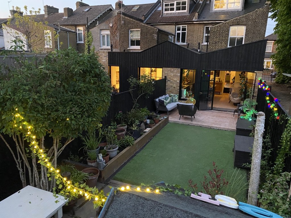 A backyard patio with outdoor seating, potted plants, string lights, and a small grass area, surrounded by a black fence and neighboring brick houses in the background.