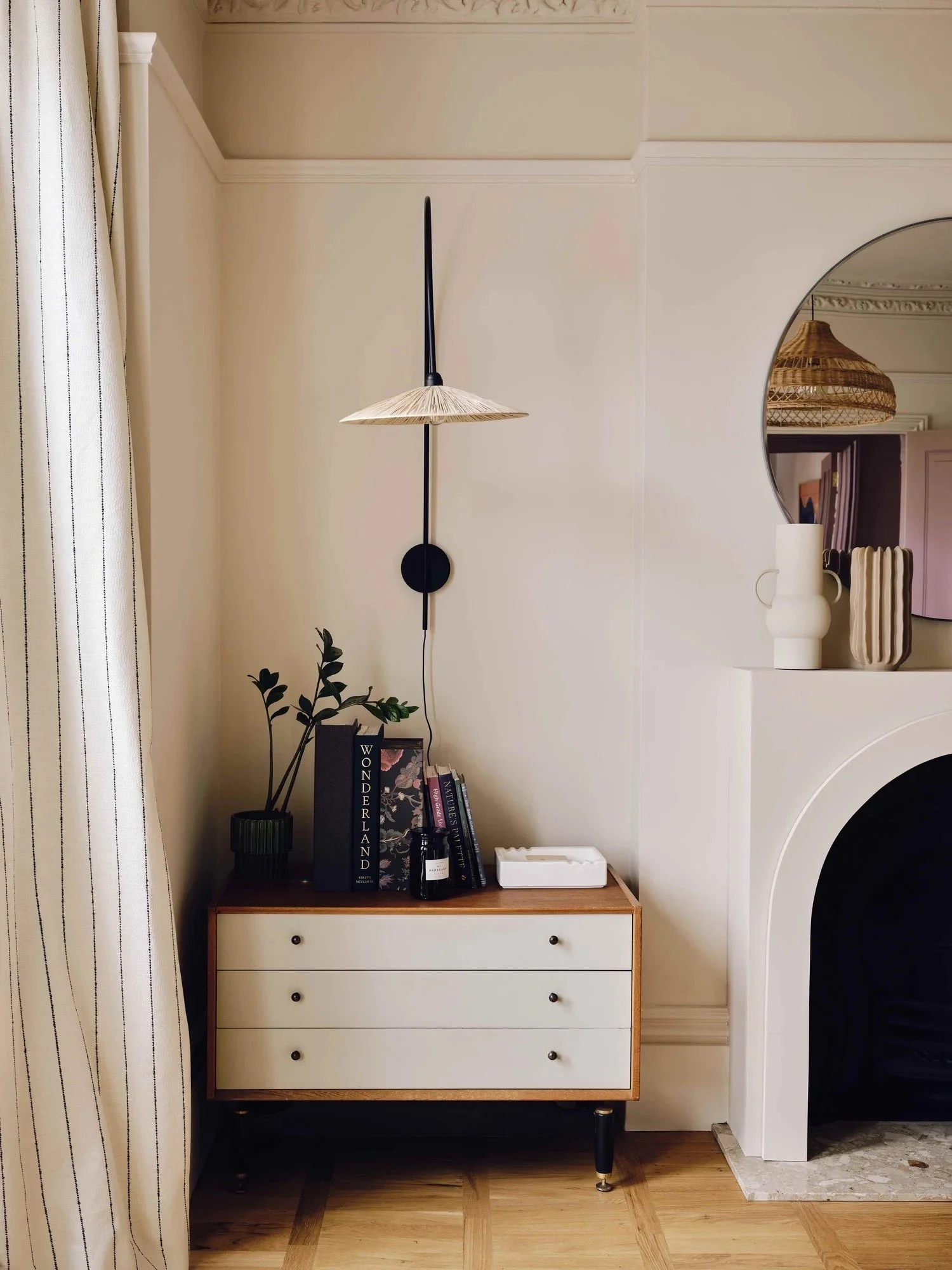 A mid-century modern dresser with white drawers and a wooden top, topped with books, a plant, and decorative items, positioned next to a circular mirror and a fireplace with white mantel and vases, in a stylish, well-decorated room.
