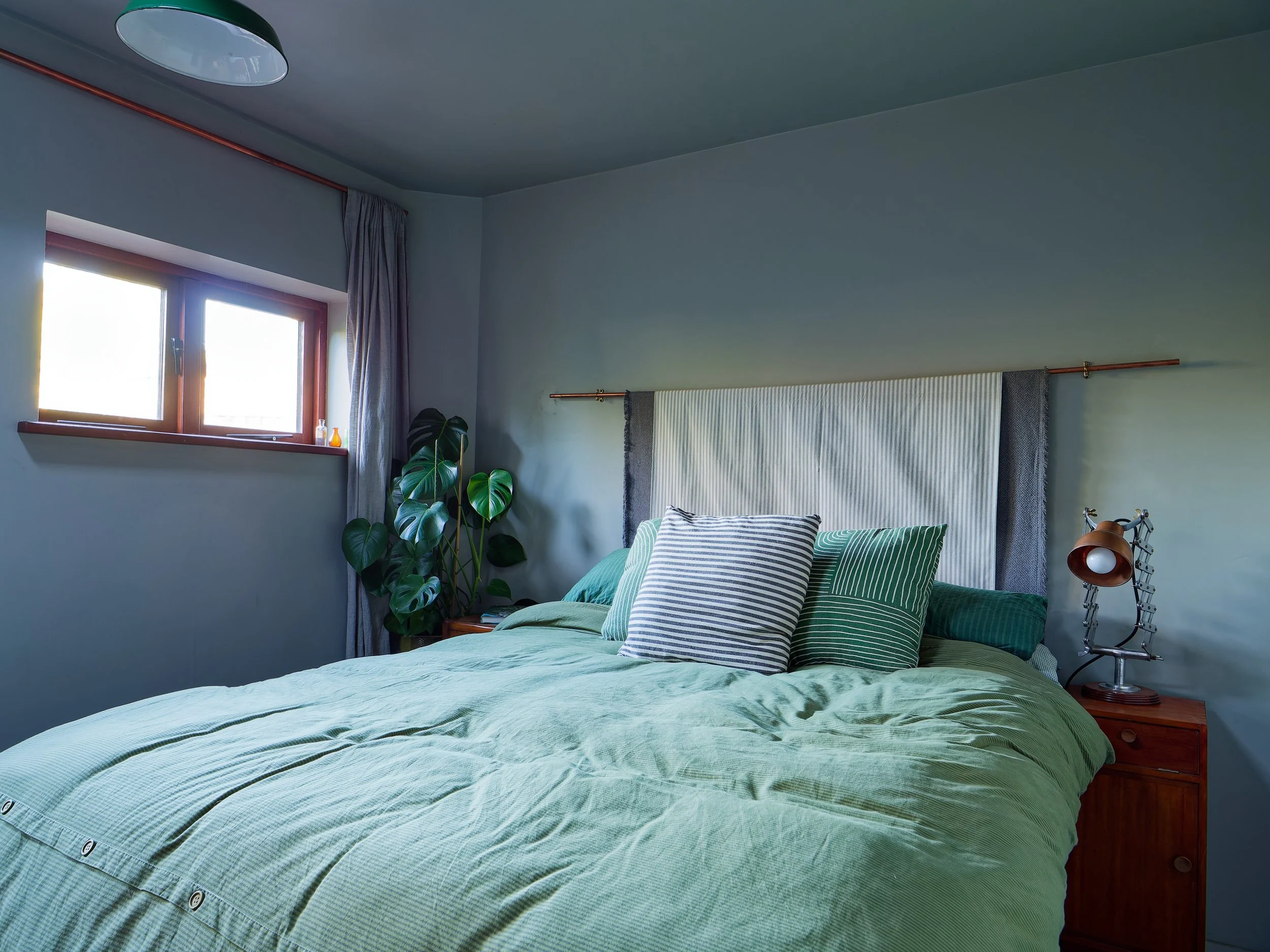 A bedroom with a bed covered in a light green quilt, multiple striped pillows, a wooden nightstand with a copper-colored lamp, a window with curtains, and a large leafy plant in the corner.
