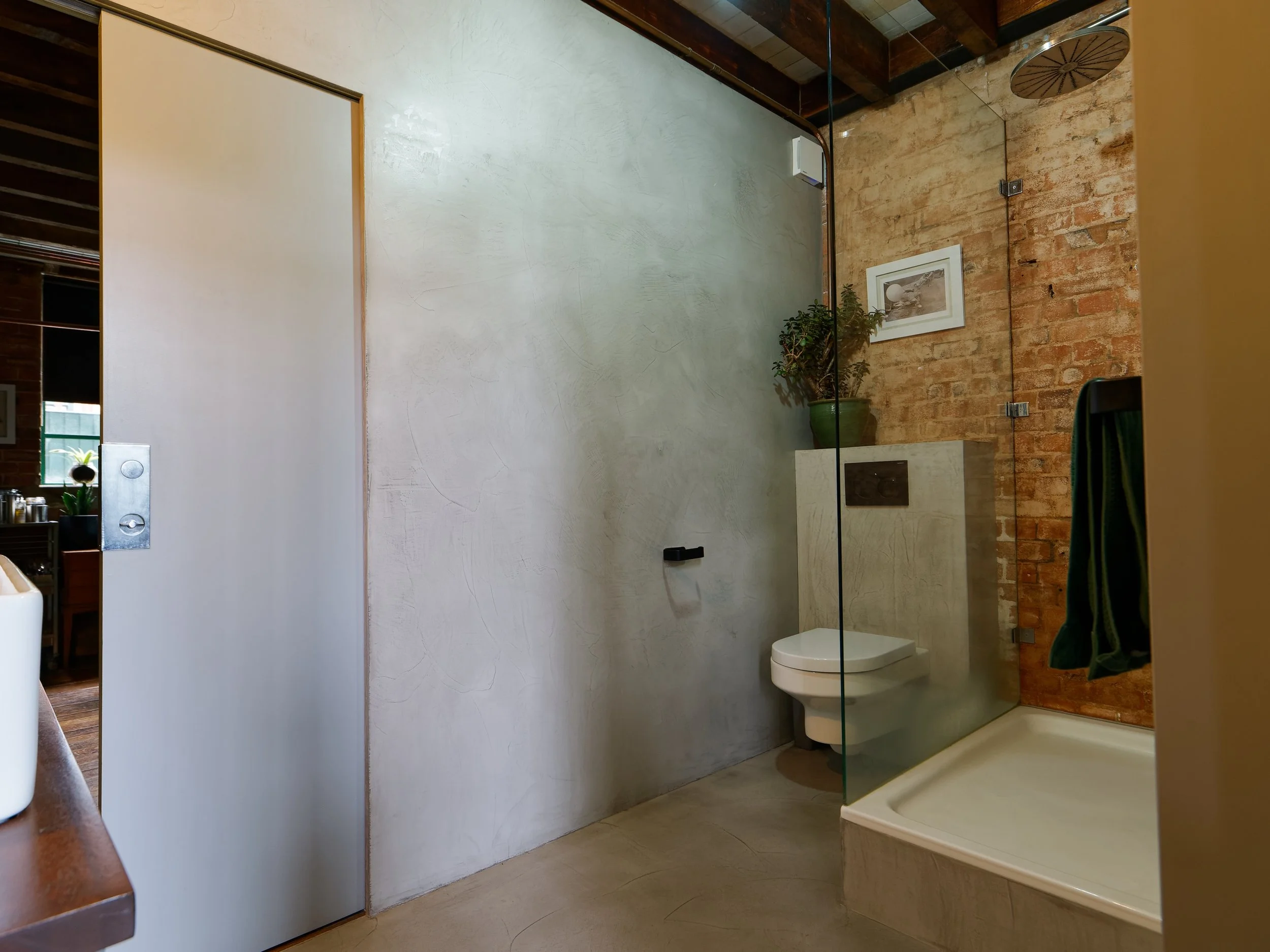 Bathroom with a brick wall shower area, ceramic toilet mounted on a concrete platform, a glass panel, a potted plant, and a framed picture on the wall.