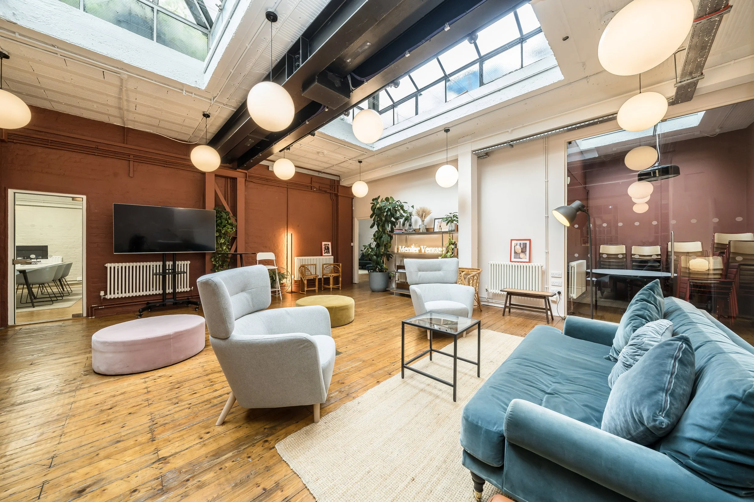 Modern living room with wooden floors, a blue sofa, white armchairs, a glass coffee table, and a wall-mounted TV. Large skylights illuminate the space, which is decorated with plants and contemporary lighting fixtures.