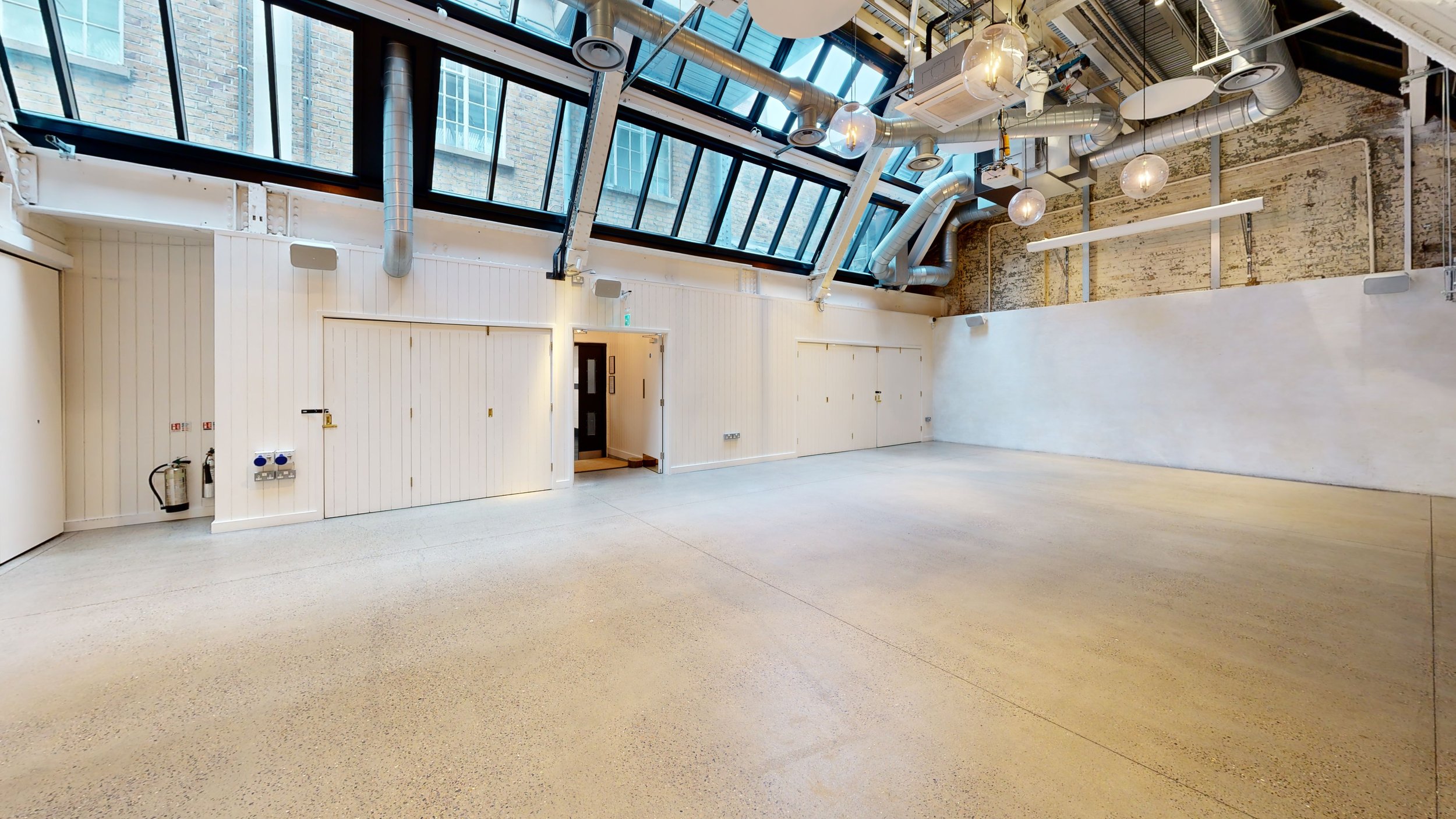 Empty industrial-style room with polished concrete floor, white paneled walls, large windows, exposed ducts, hanging light fixtures, and a small doorway.