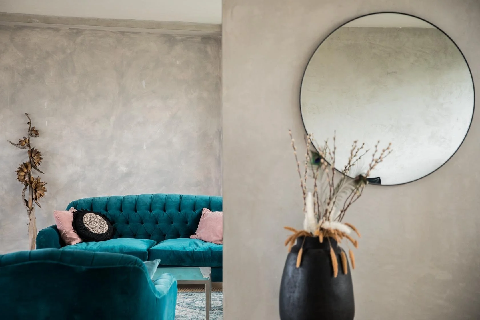 A living room with a turquoise tufted sofa decorated with pink and black pillows, a glass side table, a tall dried flower arrangement to the left, and a round wall mirror reflecting the wall behind it. The wall is textured and painted in a neutral to