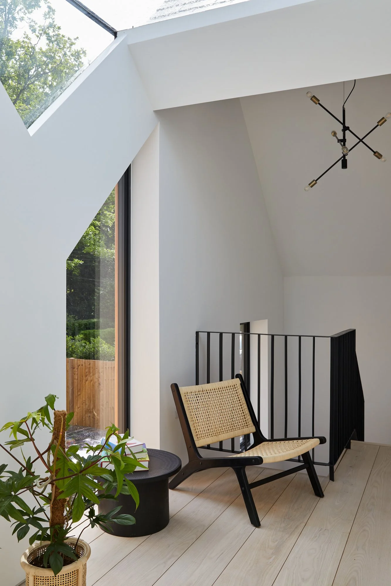 A modern interior space with a slanted ceiling and large window, featuring a black and beige woven chair, a small black round table with books and a plant, and a black railing.
