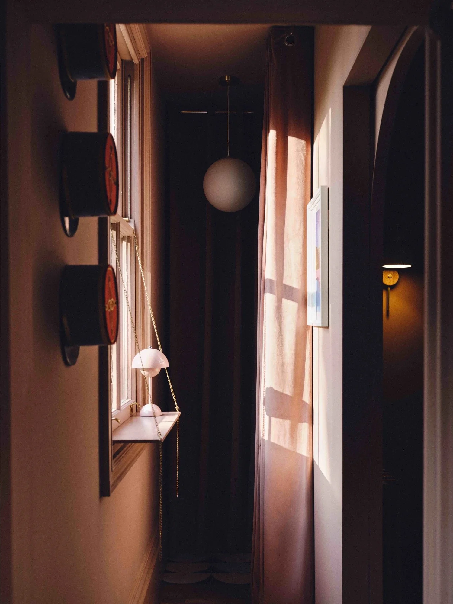 A cozy interior hallway with natural light streaming through a window with sheer curtains, a hanging spherical ceiling light, a small white shelf with a lamp in front of the window, and wall-mounted cylindrical decorations on the left.