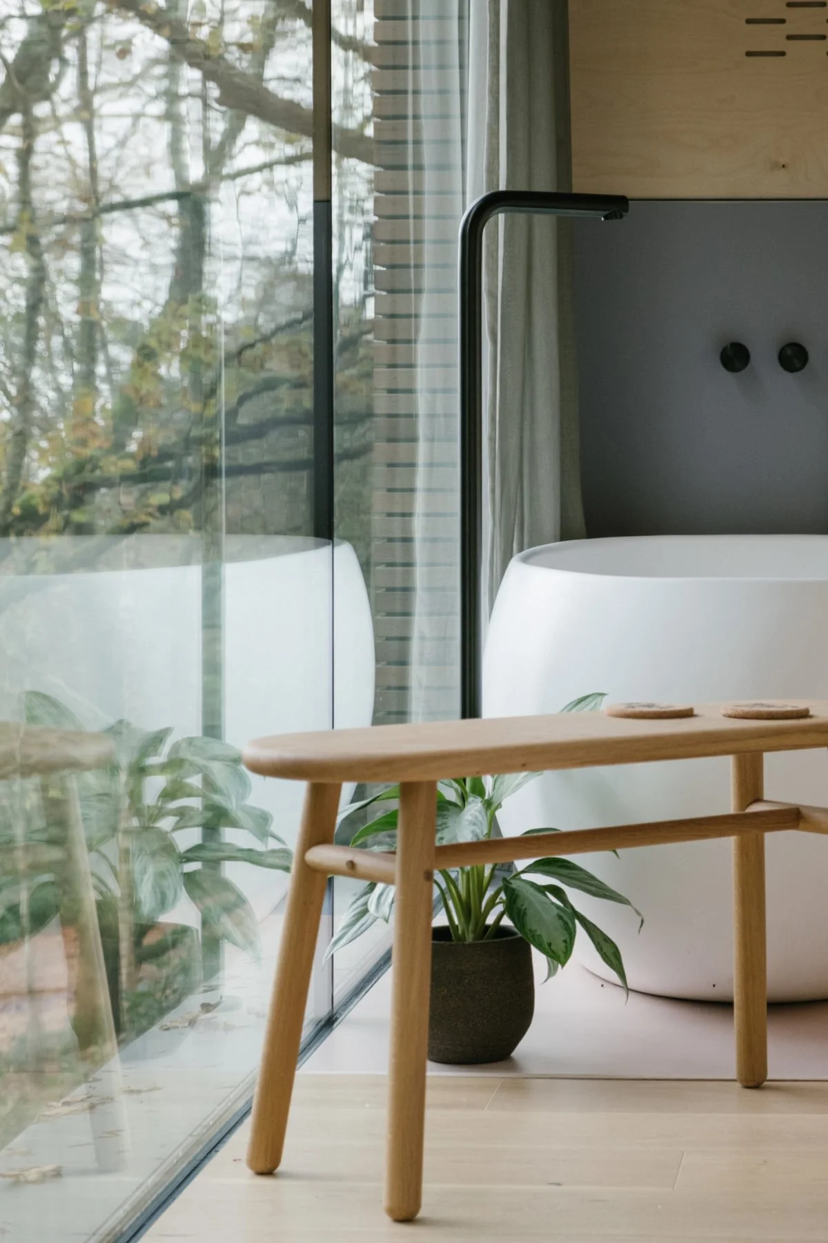 Modern bathroom scene with a black floor lamp, potted plant, white bathtub, wooden bench, large glass window showing trees outside, and beige background.