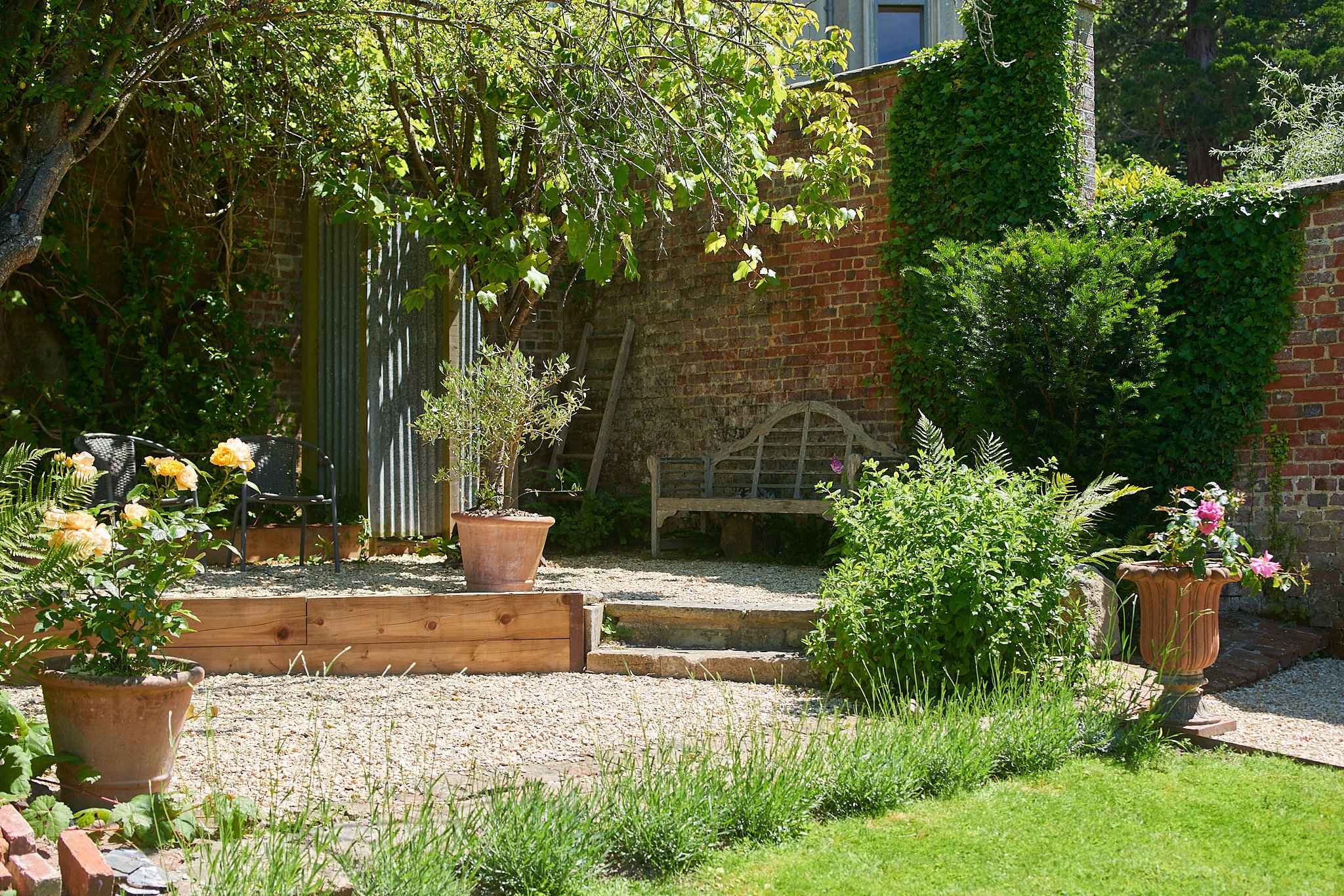 A small, sunny garden with potted plants, a stone bench, two black chairs, lush green bushes, and brick walls, with a tree providing shade.