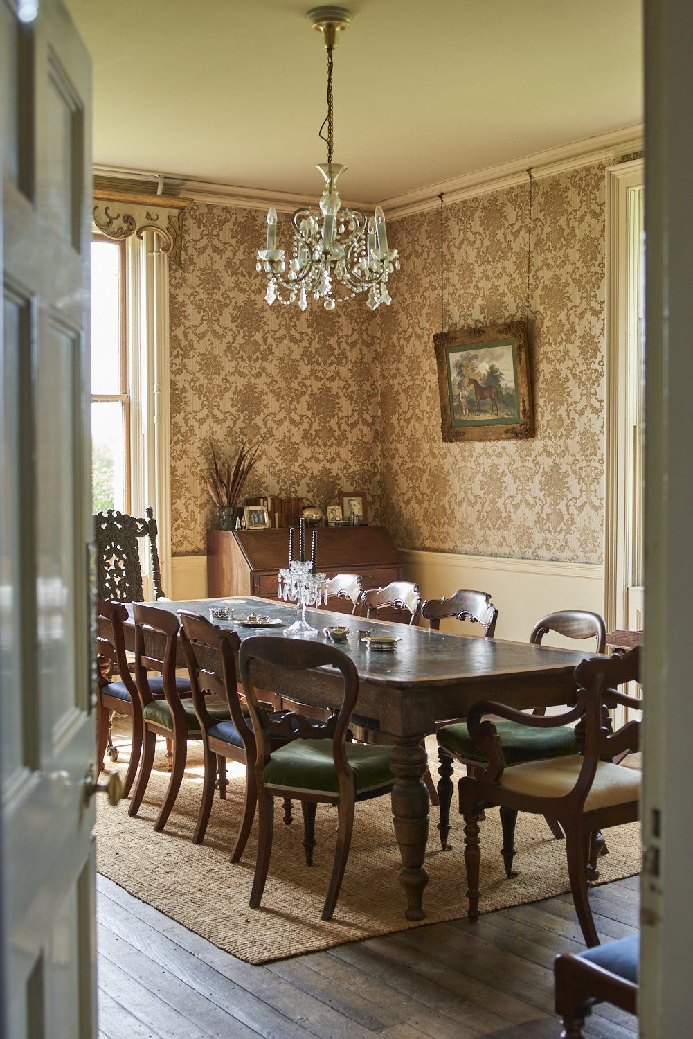 A vintage dining room with a dark wood table and ten matching chairs, chandelier hanging from the ceiling, wallpapered walls with a framed picture, a wooden sideboard with framed photos, and sunlight coming through tall windows.