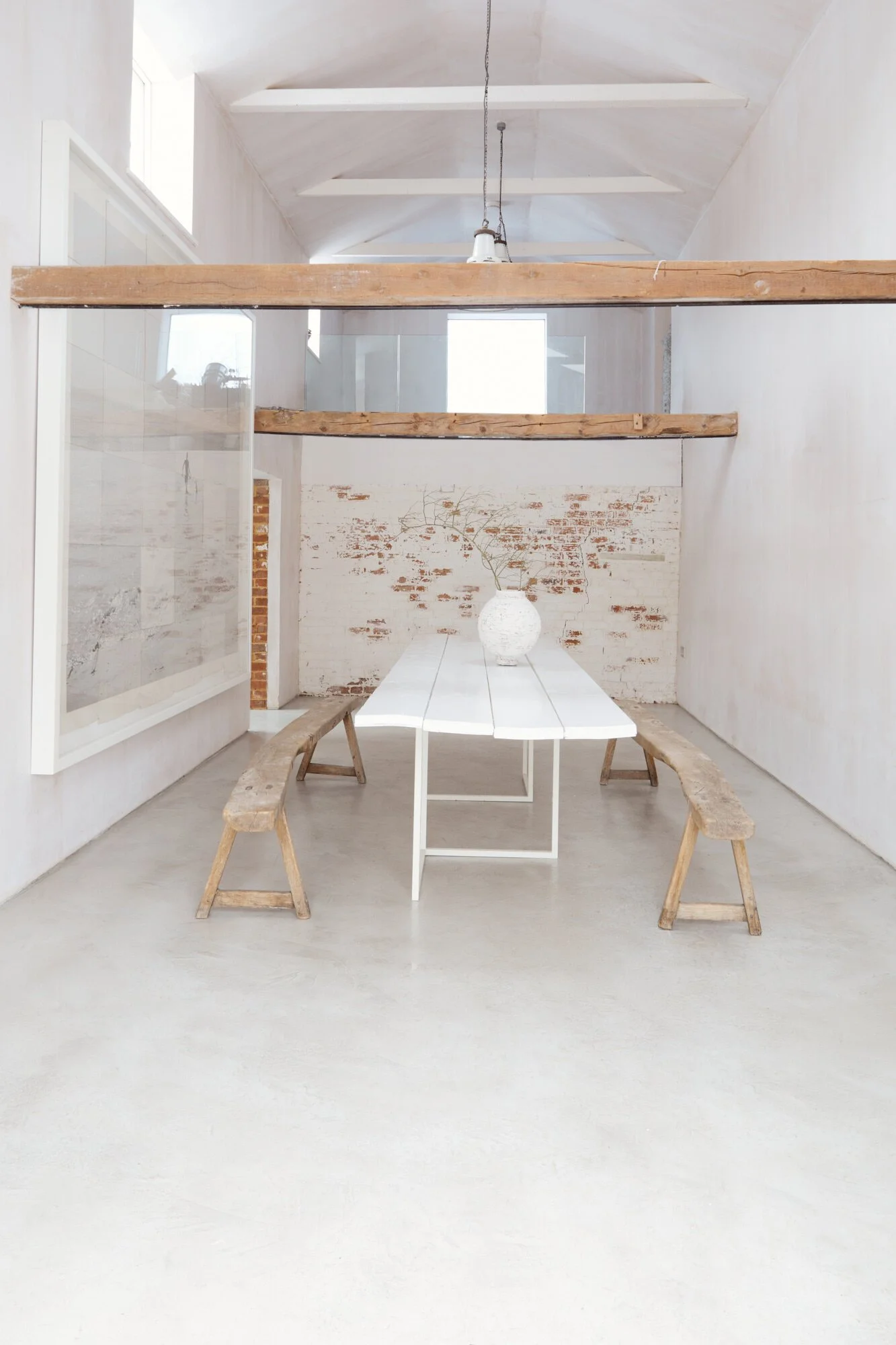 Minimalist dining area with a white table, wooden benches, a white vase with branch, brick wall in background, high ceiling, exposed beams, window, and hanging light fixture.