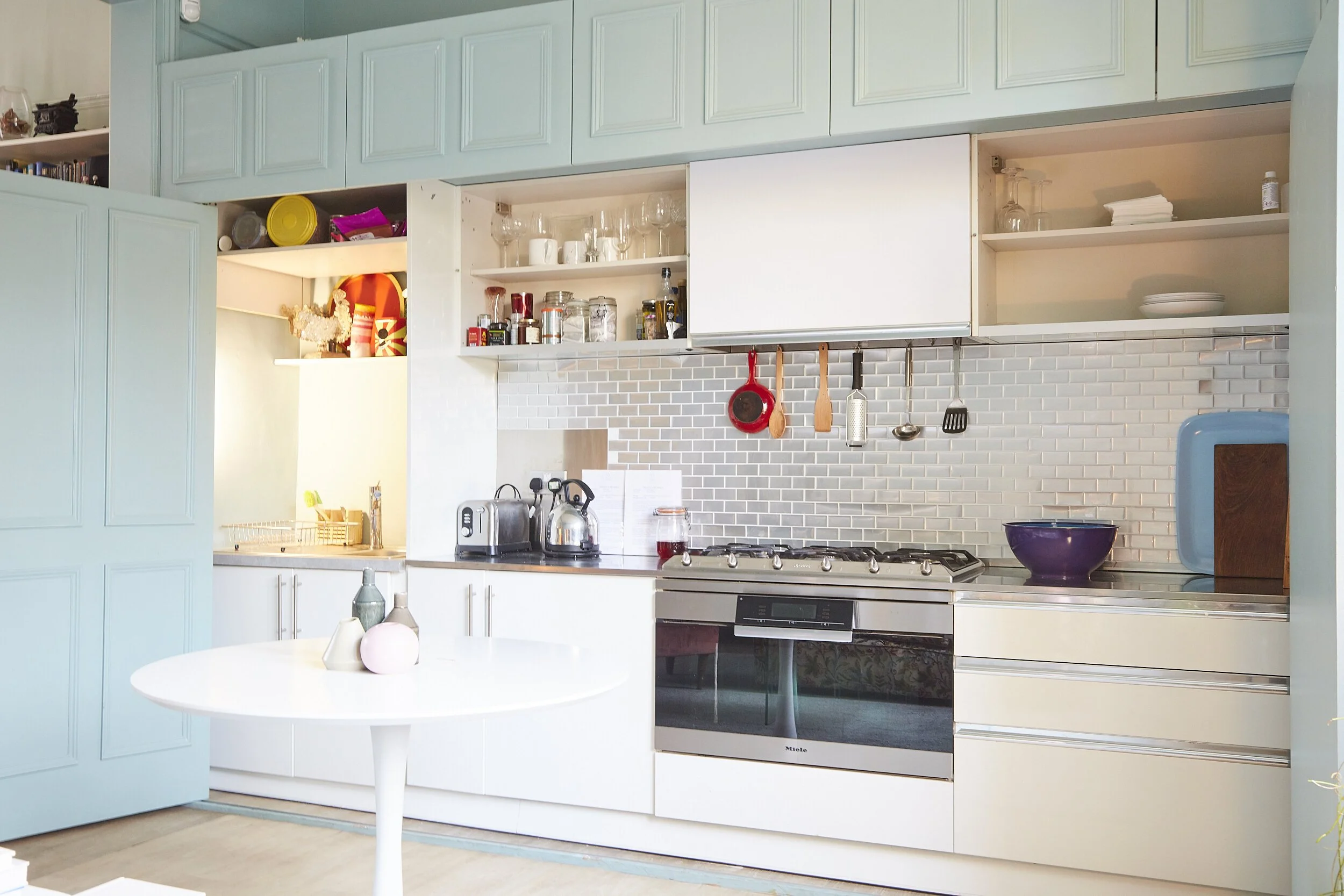 Modern kitchen with white cabinetry, a stainless steel oven, a light blue cabinet door, hanging pots and utensils, and a white round table with vases.