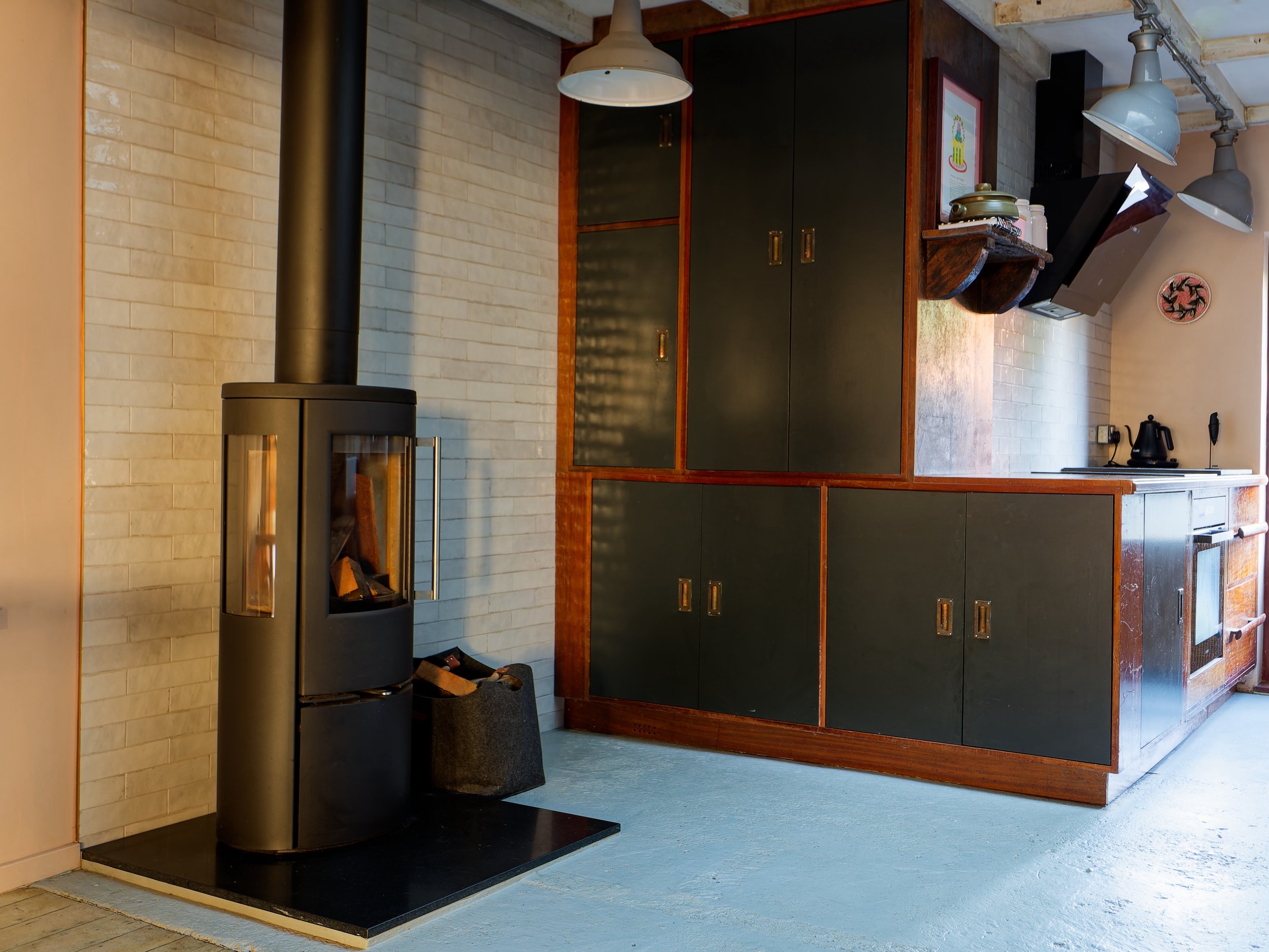 A modern kitchen with a wood and black cabinet, a black wood stove with a chimney, and some kitchen items on open shelves. Soft natural light illuminates the space.