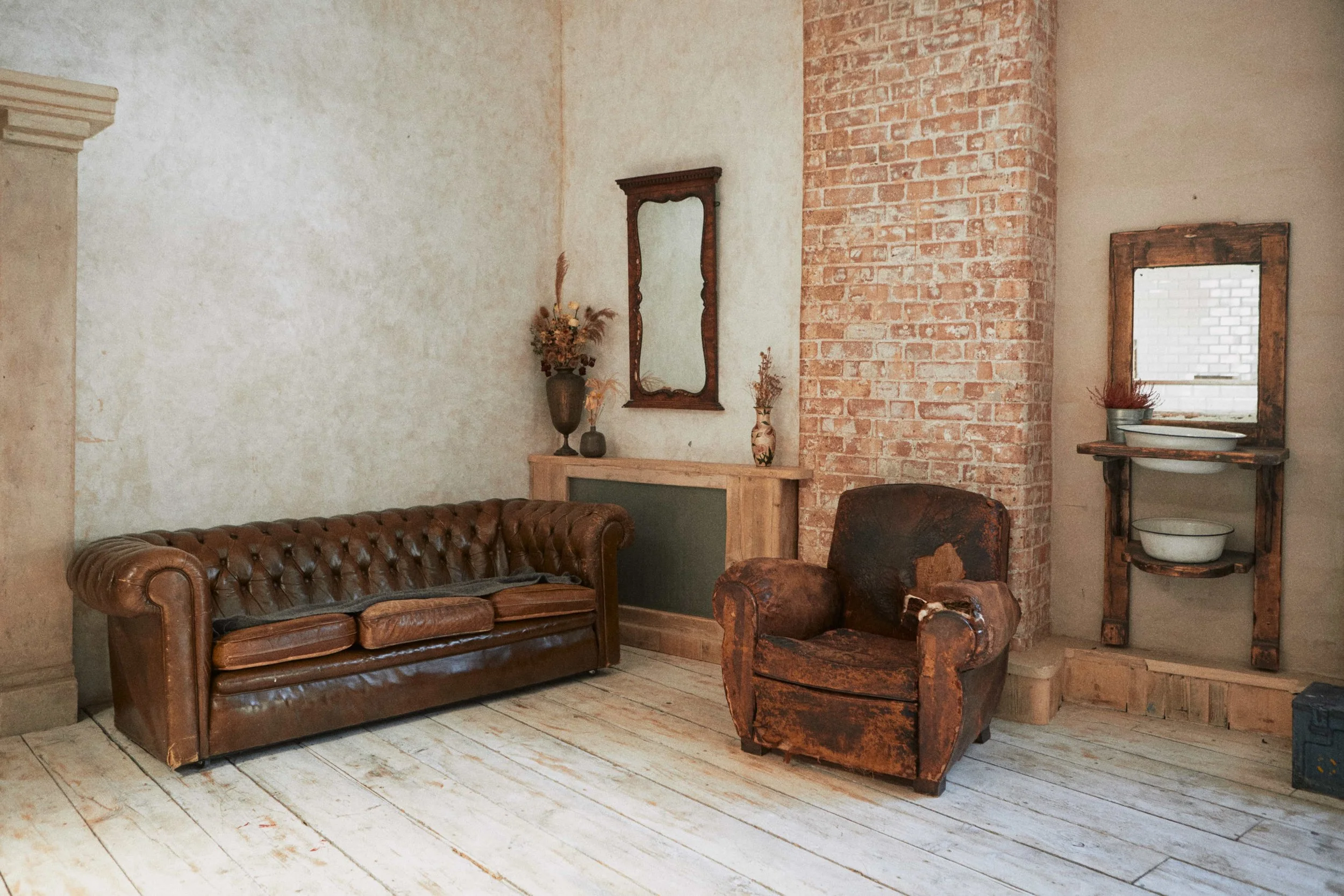 A rustic living room with a worn leather sofa, a distressed armchair, vintage mirror, and shelving with dishes, against a brick and beige wall.