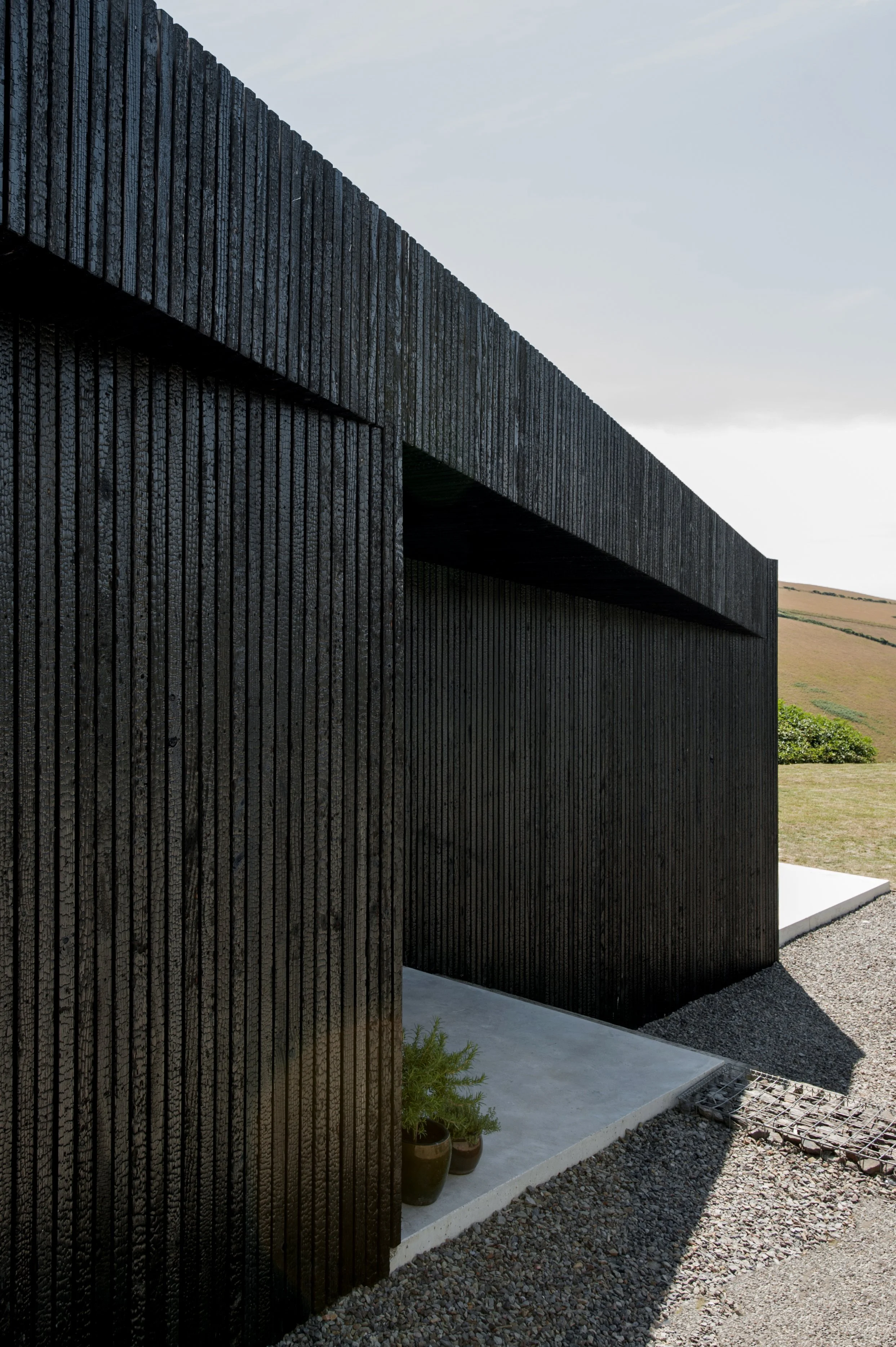 Close-up of a modern building with black vertical wooden slats, overlooking a grassy landscape with rolling hills in the background.