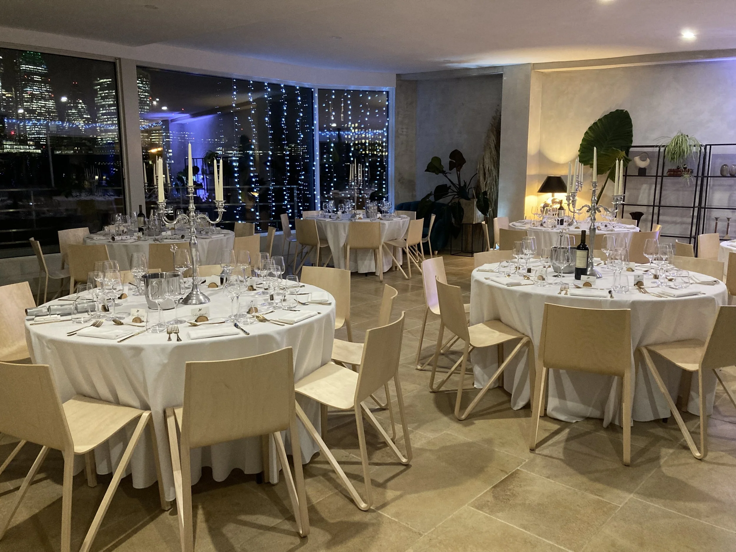 Elegant dining room set for a formal event with white tablecloths, wooden chairs, silver candelabras, and glassware, with city skyline and string lights visible through large windows at night.