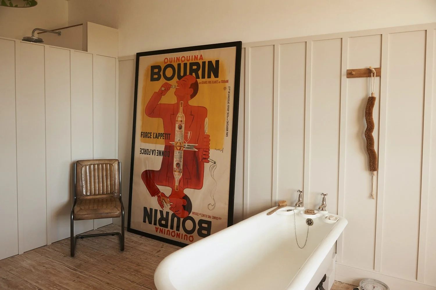 A vintage-style bathroom corner featuring a white bathtub, a brown cushioned chair, a poster leaning against the wall, and a wooden wall decoration. The background has white paneled walls.