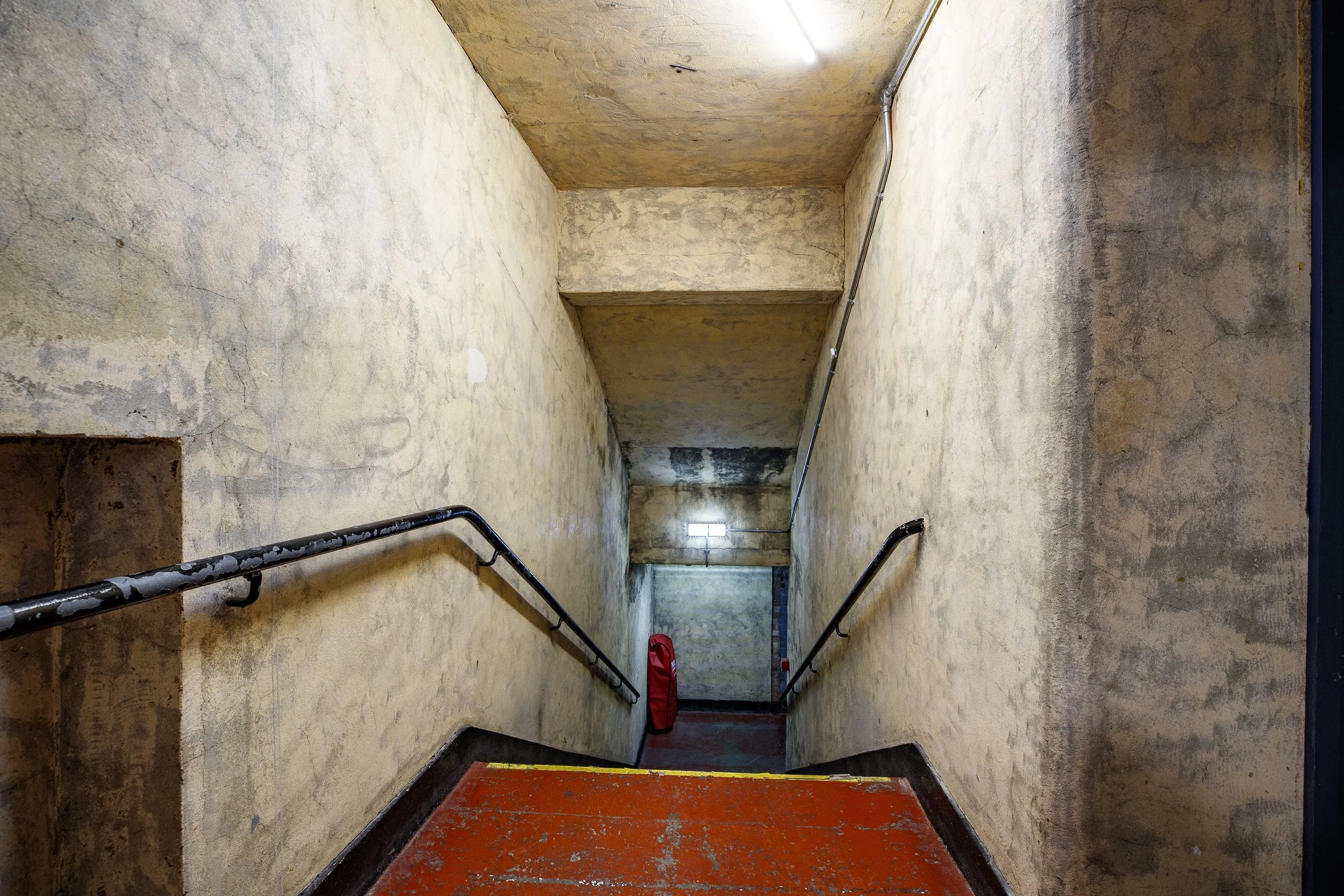 An indoor stairwell with beige stained walls, black handrails on each side, a red object at the bottom, and a fluorescent light near the ceiling illuminating the area.