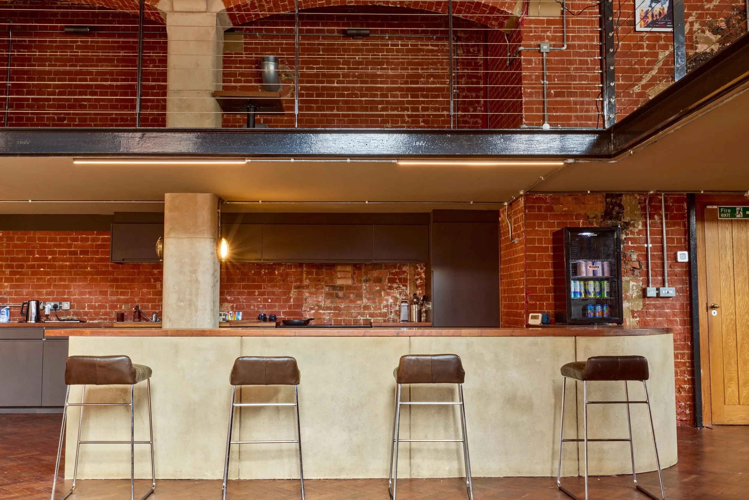 Modern kitchen and bar area with exposed red brick walls, a concrete column, and four brown leather bar stools in front of a light-colored curved counter. There is a shelving unit with canned beverages on the right and a wooden door with an emergency
