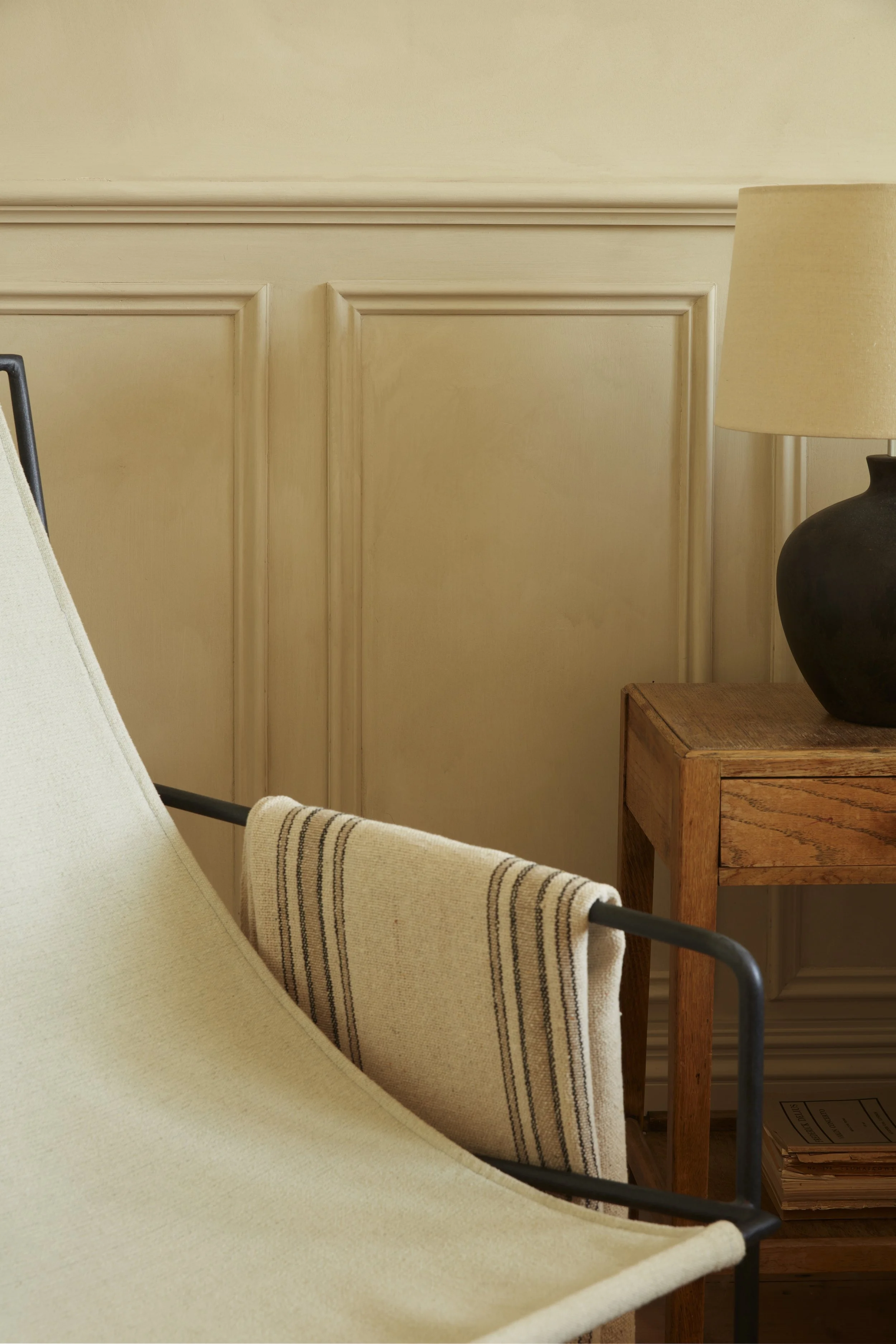 A cozy corner with beige walls, a beige hammock, a wooden side table, a black ceramic lamp with a beige lampshade, and a folded striped blanket.
