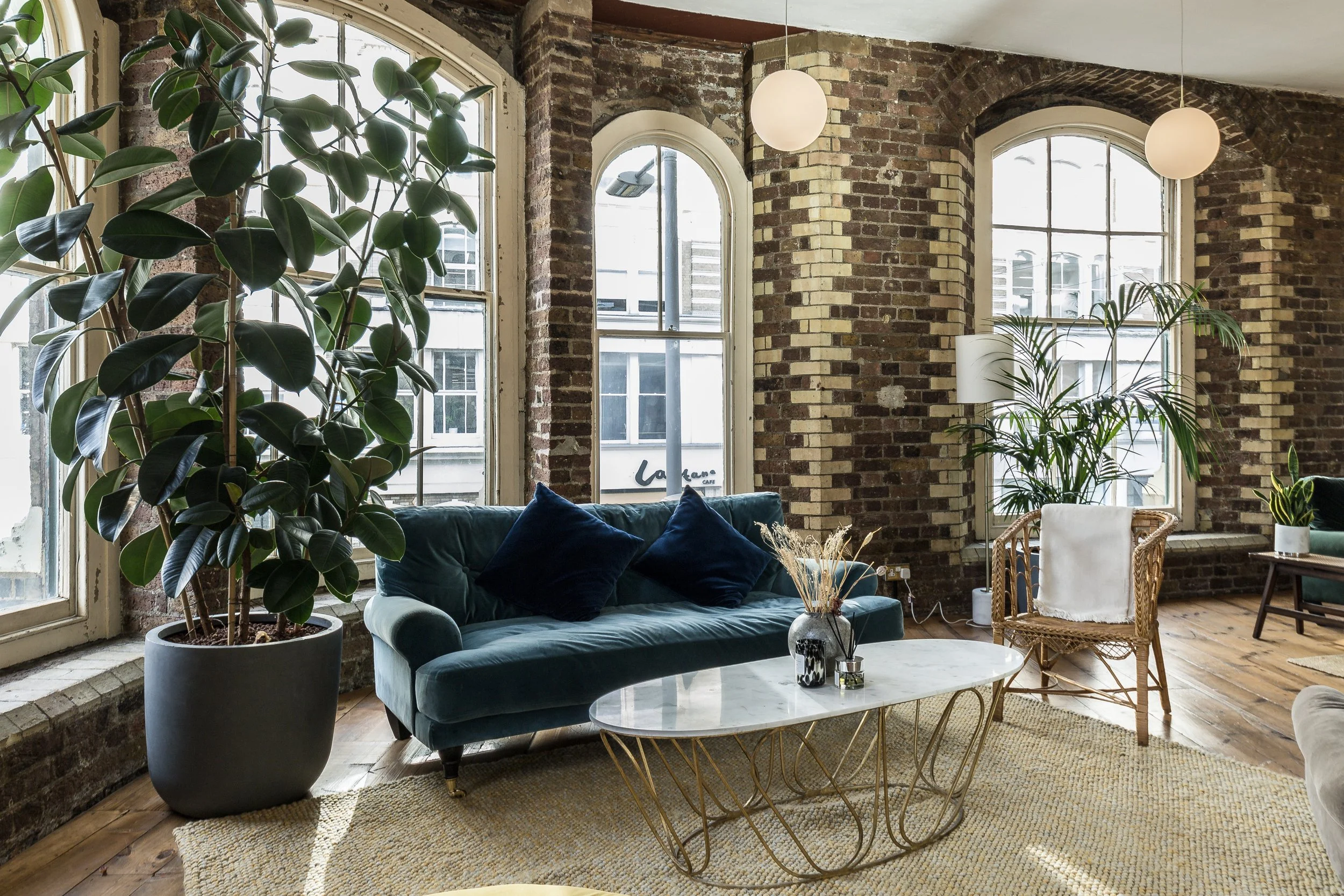 Living room with large windows, exposed brick walls, a blue velvet sofa with dark cushions, round coffee table with decorative vases, wicker chair with white pillow, tall indoor plants, and a light-colored rug.