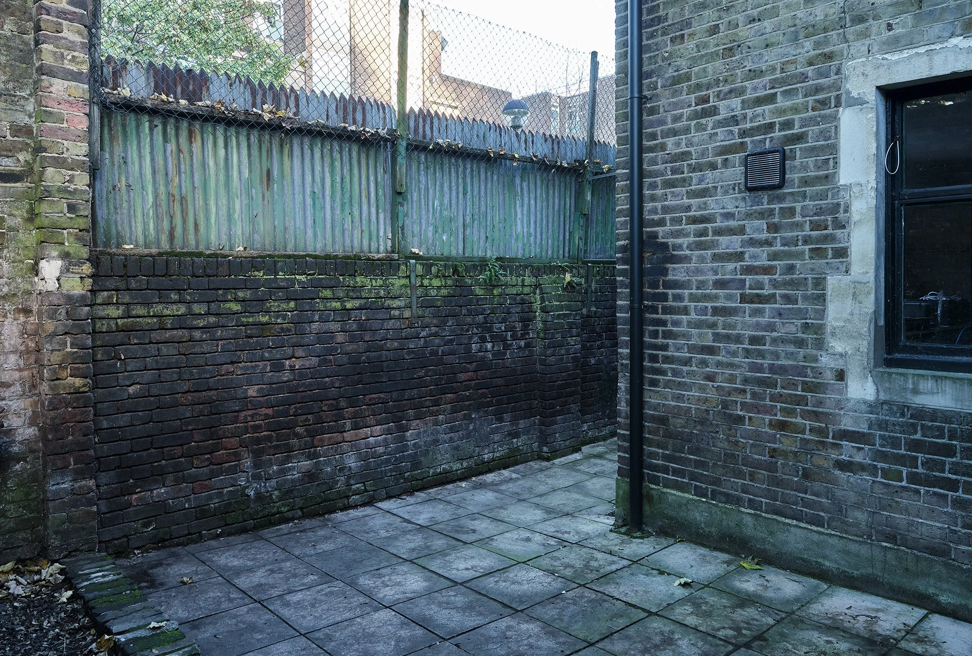 An outdoor courtyard area enclosed by brick and metal fences, with a concrete paver floor and a window on a brick building wall.