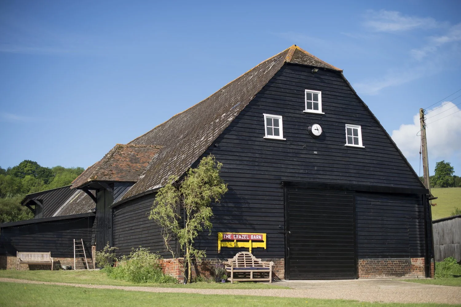 A large black barn with a weathered shingled roof, four windows, and a door, with a sign that reads "The Crazel Barn" in front. There is a bench, a small tree, and some greenery near the barn, with a blue sky and green hills in the background.