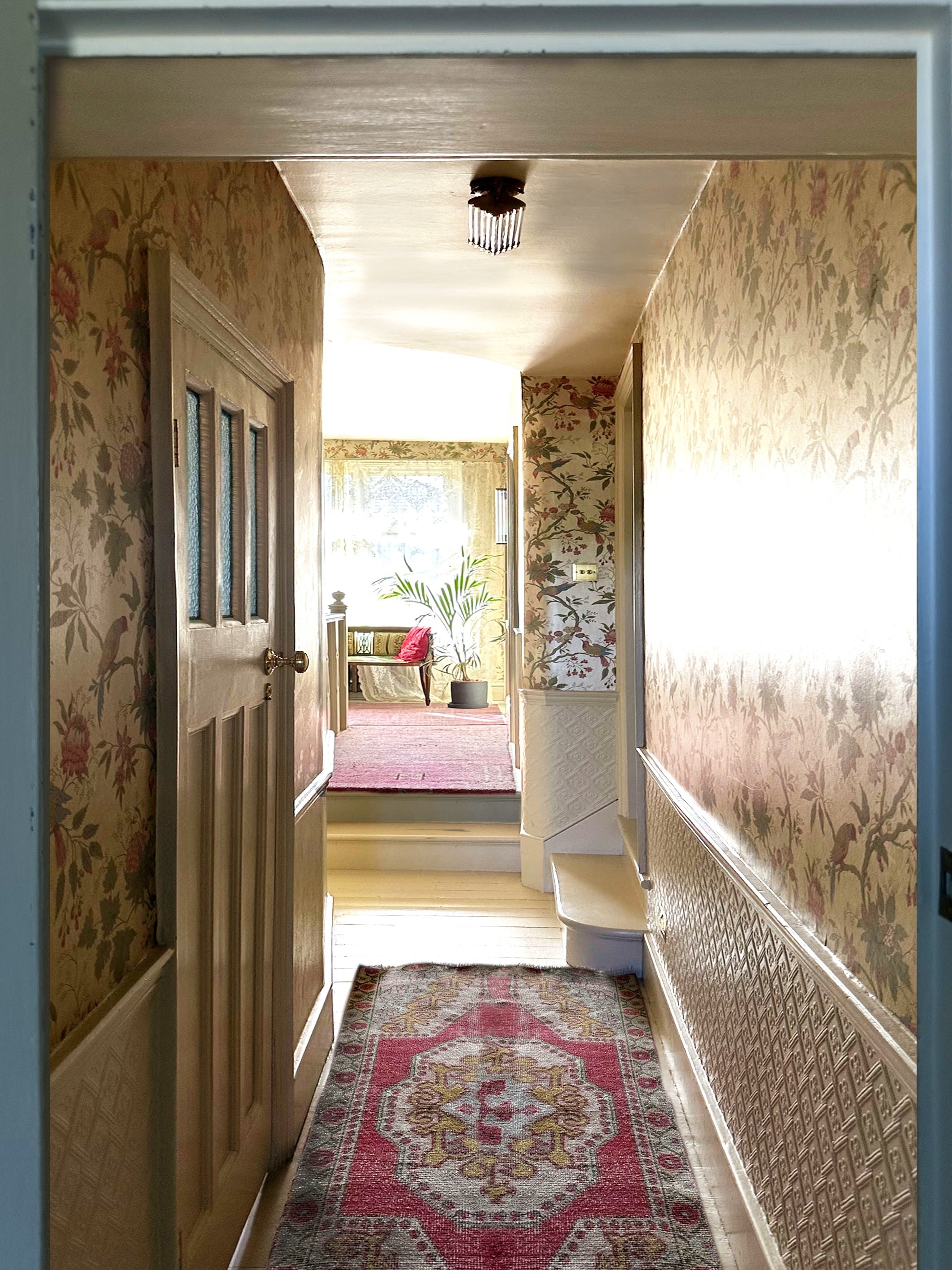 A hallway leading to a sunlit room with a window, a green plant, and a red cushion on a bench, with floral wallpaper and a colorful rug.