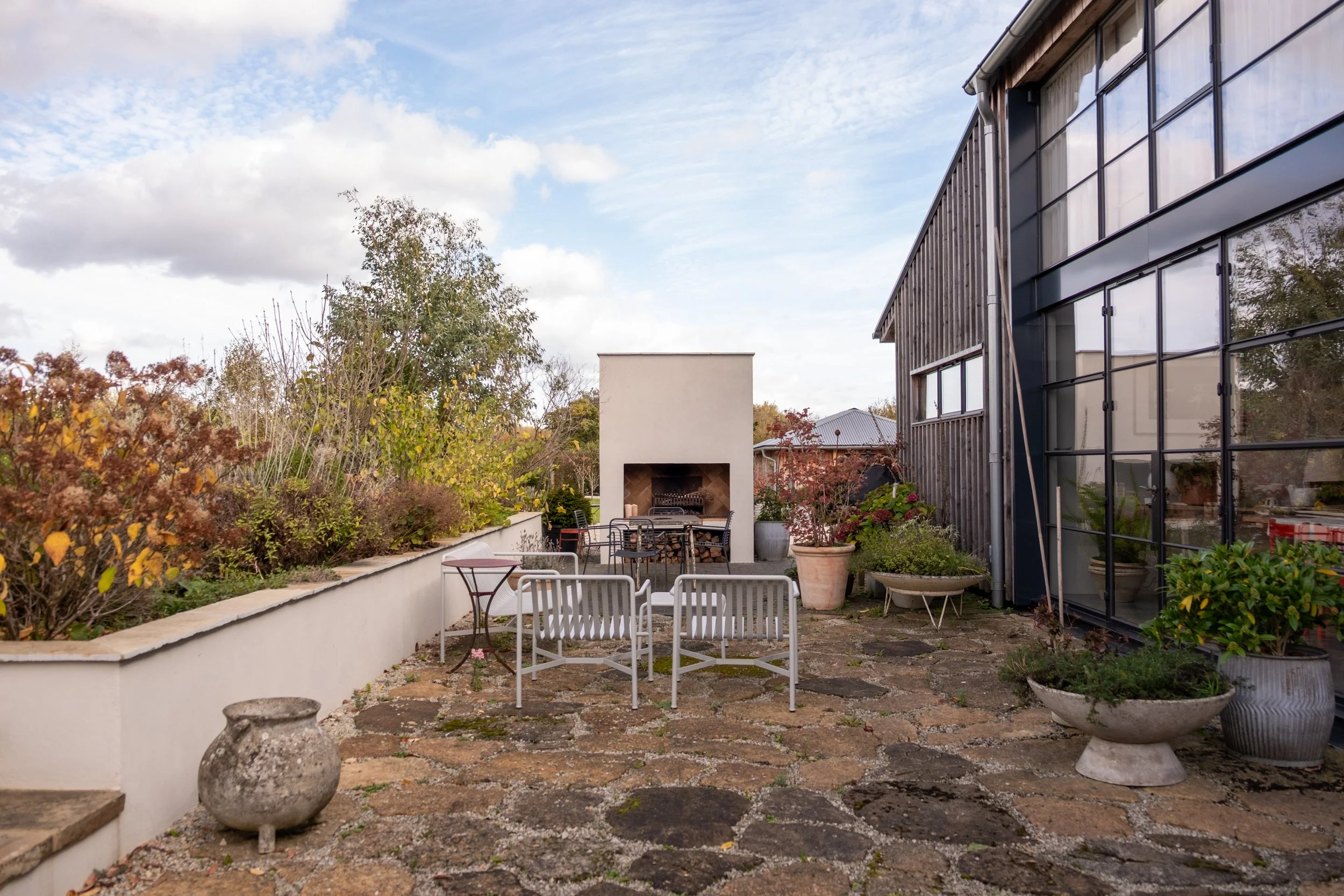 Outdoor patio area with chairs, potted plants, a white wall with a built-in fireplace, trees, and a modern building with large glass windows on a partly cloudy sky day.