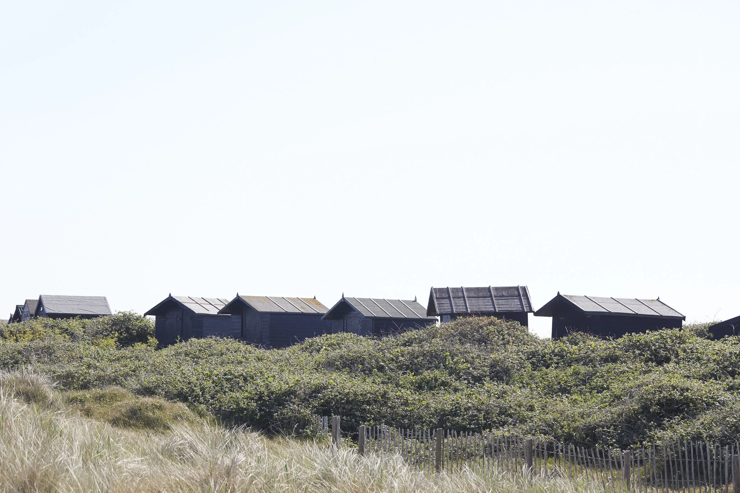 Several small, dark-colored wooden houses or sheds on a hill covered with bushes and tall grass below, with a clear bright sky above.