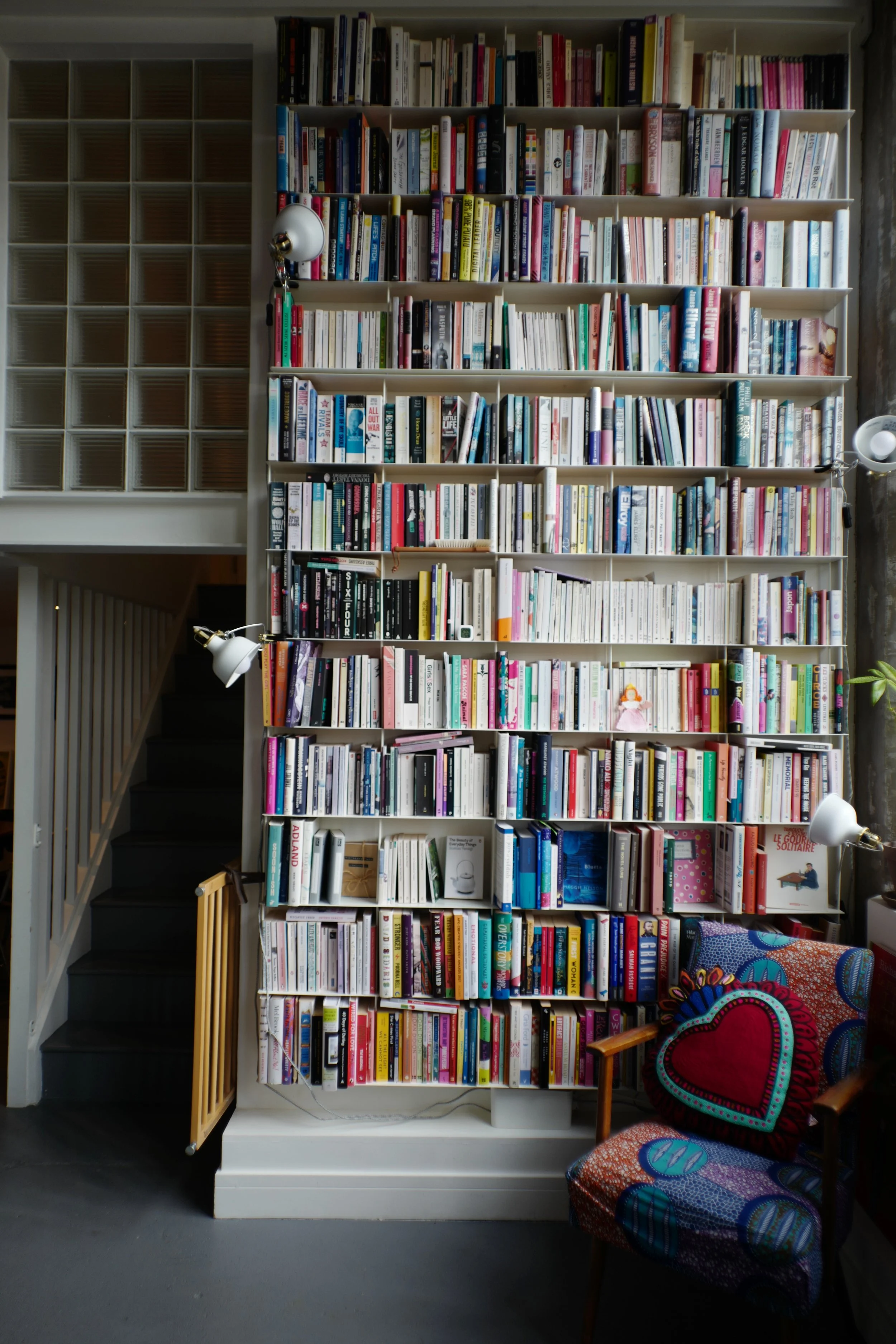 A tall bookshelf filled with a variety of books in a room with a staircase to the left and a colorful patterned armchair with a heart-shaped pillow to the right.