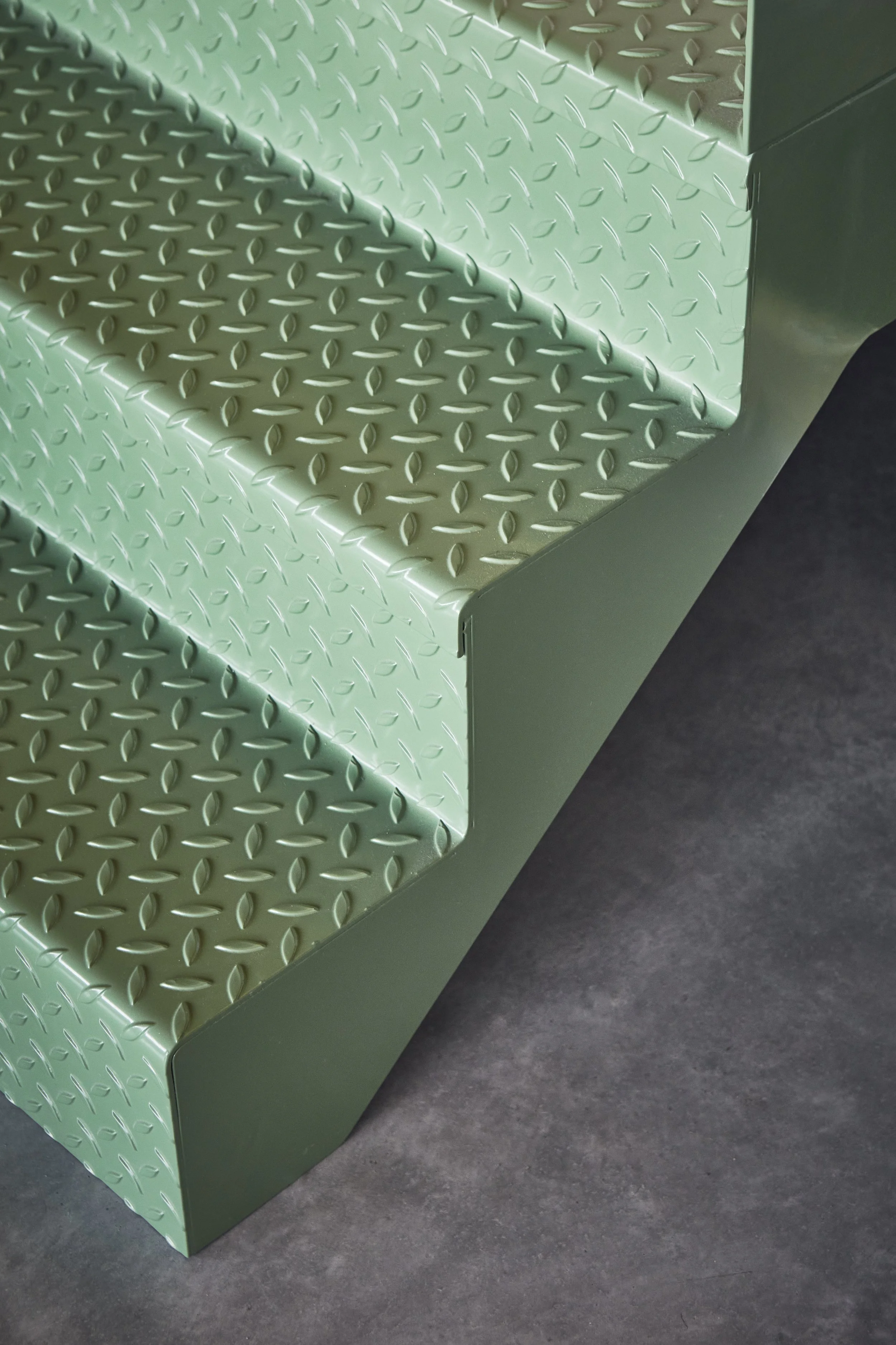 Close-up of green metal stairs with a diamond plate pattern, positioned against a dark floor.