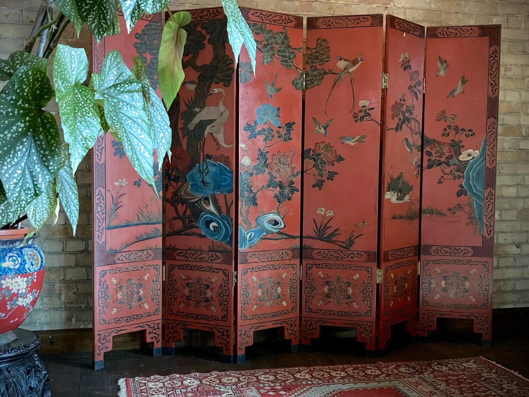 Decorative Asian folding screen with red background and painted cranes, flowers, and natural scenery, positioned against a brick wall in front of a patterned rug.