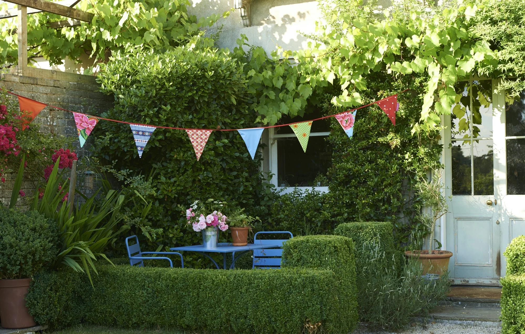 A cozy backyard scene with a blue metal table and two matching chairs, surrounded by neatly trimmed hedges and potted plants, with colorful bunting flags hanging overhead.