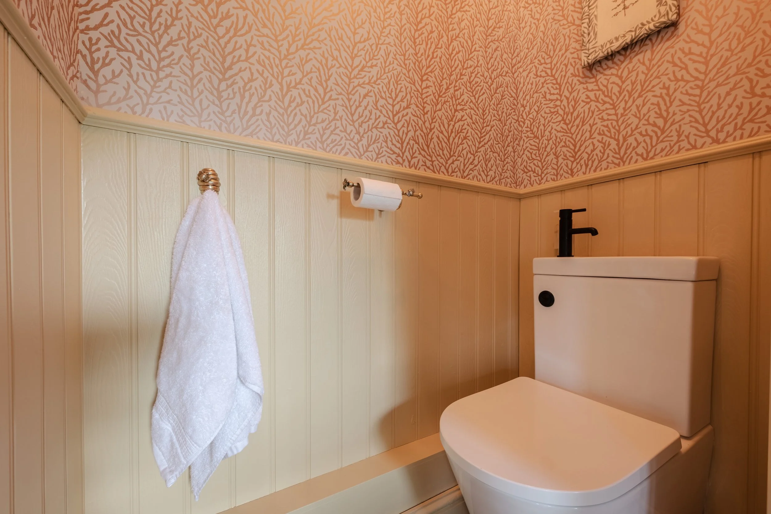 Bathroom with beige wall paneling, pink coral-patterned wallpaper, a white towel hanging on a hook, a toilet paper roll, and a modern white toilet with a black faucet.