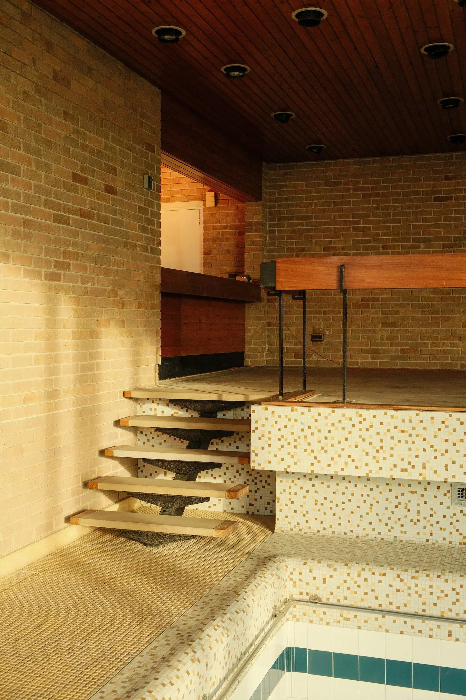 Indoor swimming pool area with tiled steps leading up to a wooden platform, brick walls, and a wooden ceiling. The pool has a tiled edge with a mosaic pattern.