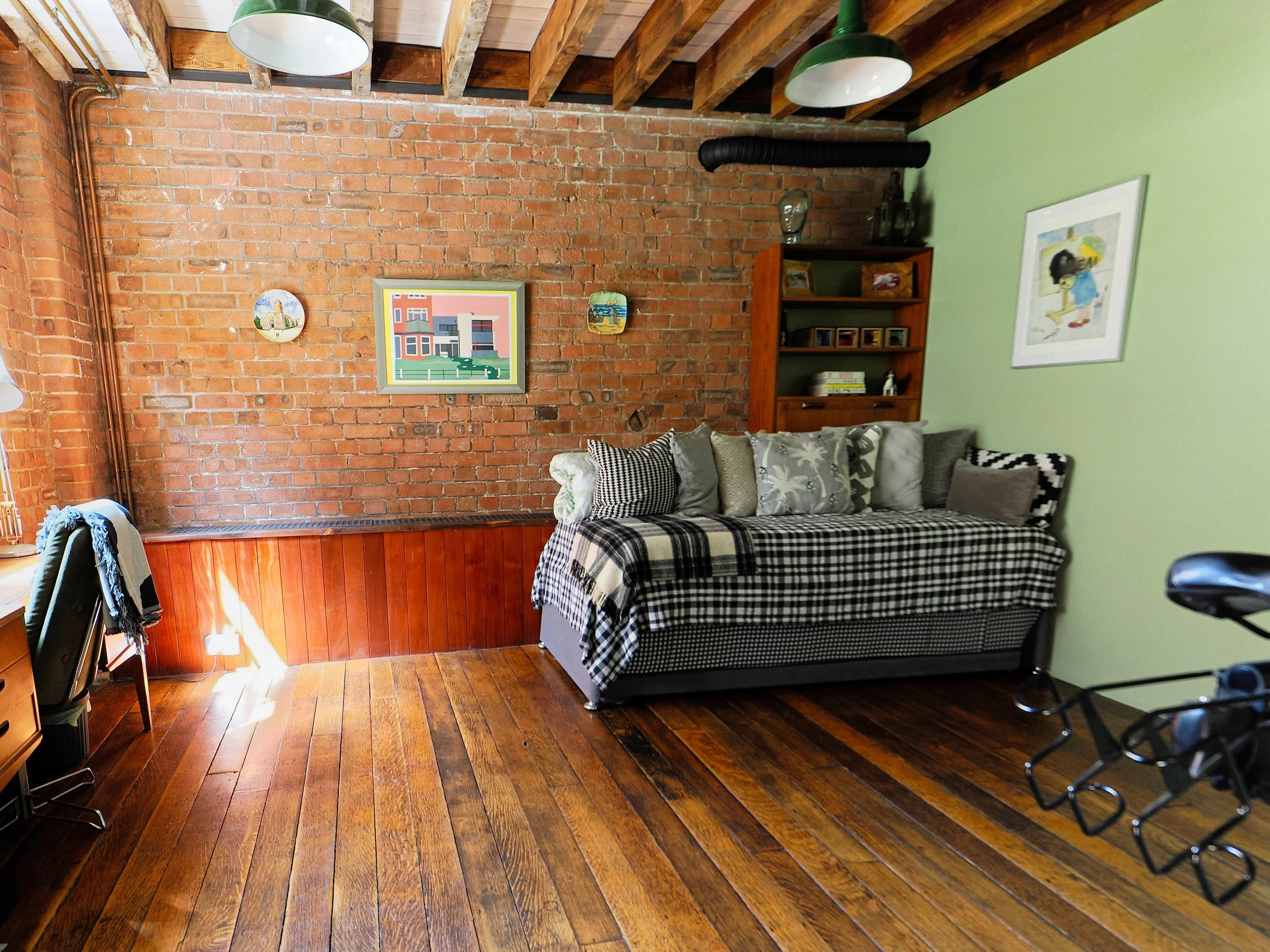 Cozy living room with exposed brick wall, wooden floor, and a green painted wall. Features a large checkerboard-patterned sofa with various cushions, a wooden bookshelf, and wall art. Part of a bicycle is visible in the foreground.
