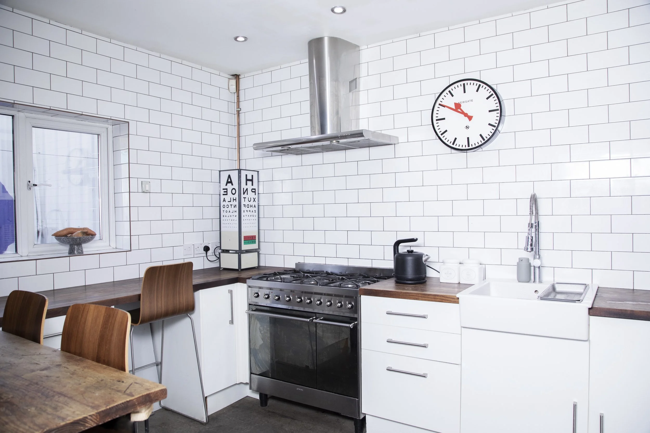 Modern kitchen with white subway tile walls, wooden countertops, a black stove, a white farmhouse sink, a black kettle, and an eye chart on a stand. There is a window with a small bowl on the windowsill and a wall clock showing about 12:45.