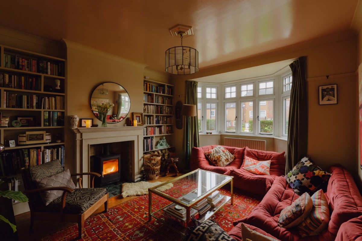 A cozy living room with a fireplace and a large bay window. It has red sofas with patterned pillows, a glass coffee table, bookshelves filled with books, a mirror above the fireplace, and various decorative items.