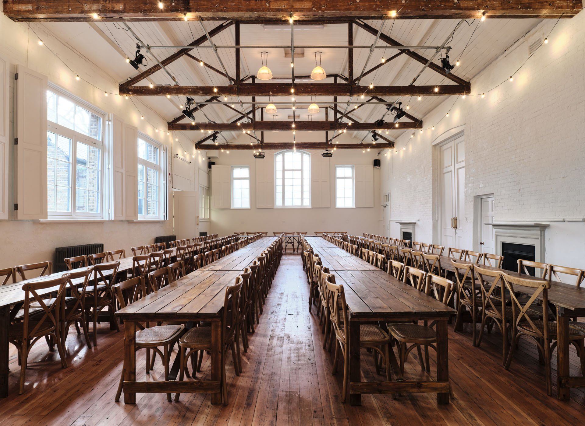 Large event space with long wooden tables and chairs arranged for a gathering, with string lights hanging from the ceiling and large windows allowing natural light.