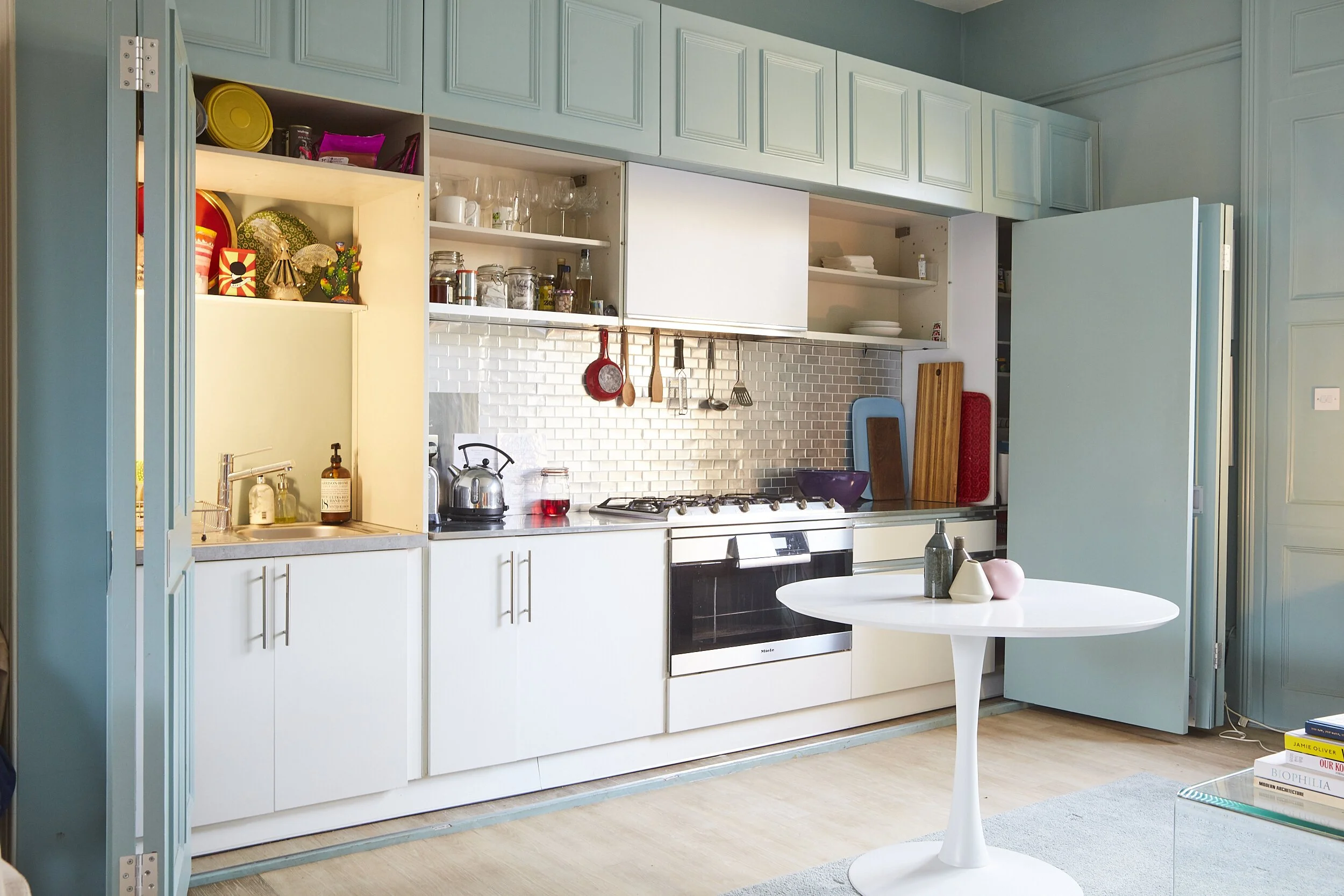Bright, modern kitchen with light blue cabinetry, white lower cabinets, a white round table with decorative items, a stainless steel stove, and open shelving with dishes, glasses, and utensils.