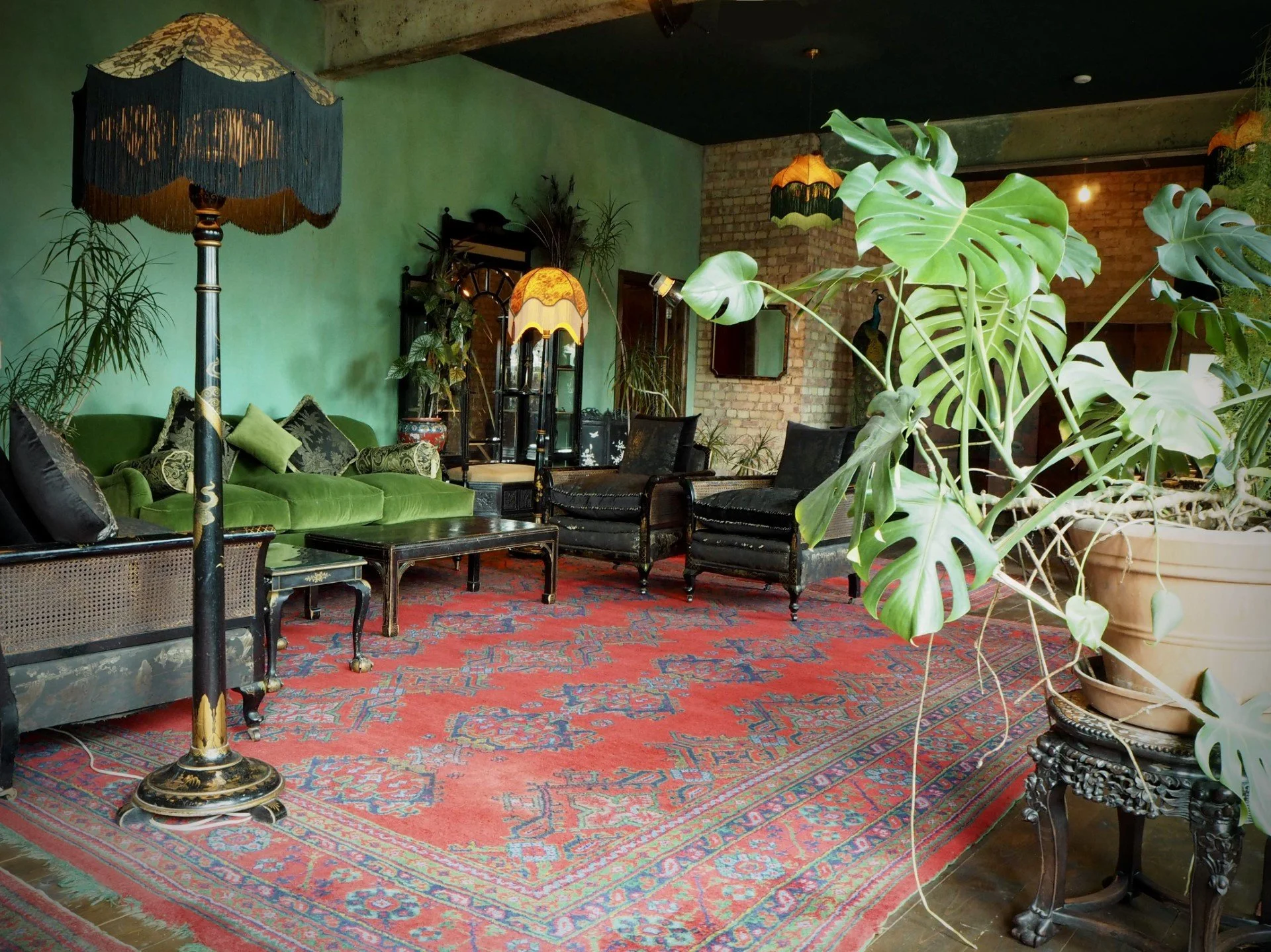 Living room with vintage style furniture, including a green velvet sofa, black armchairs, and a decorated area rug. There are multiple floor lamps with ornate lampshades, a large potted monstera plant in the foreground, and green walls with brick acc