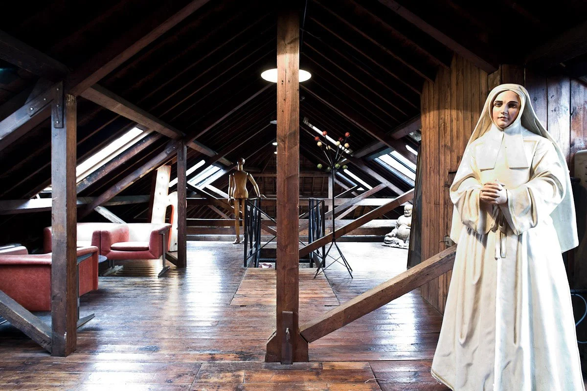 Interior of an attic with wooden beams, sunlight through skylights, a seated statue of Mother Teresa, and modern decor including red armchairs and a sculpture of an atom model.