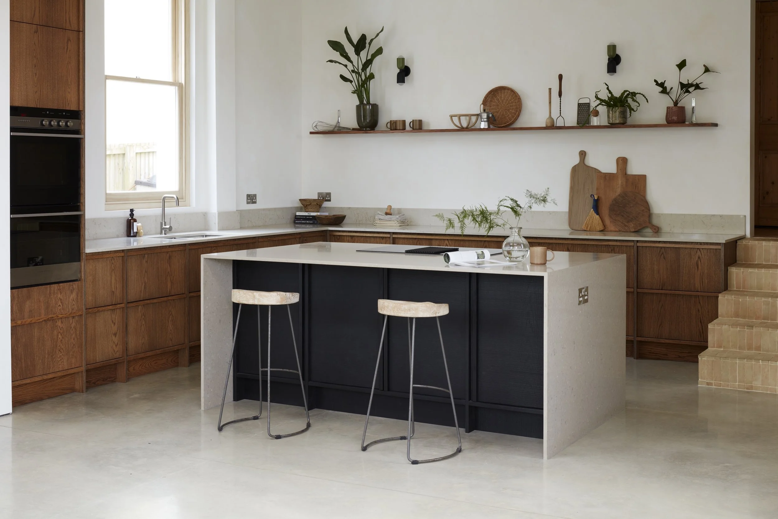 Modern kitchen with white countertops, black and wooden cabinets, open shelving with plants and kitchenware, two stools at a central island, large window letting in natural light.