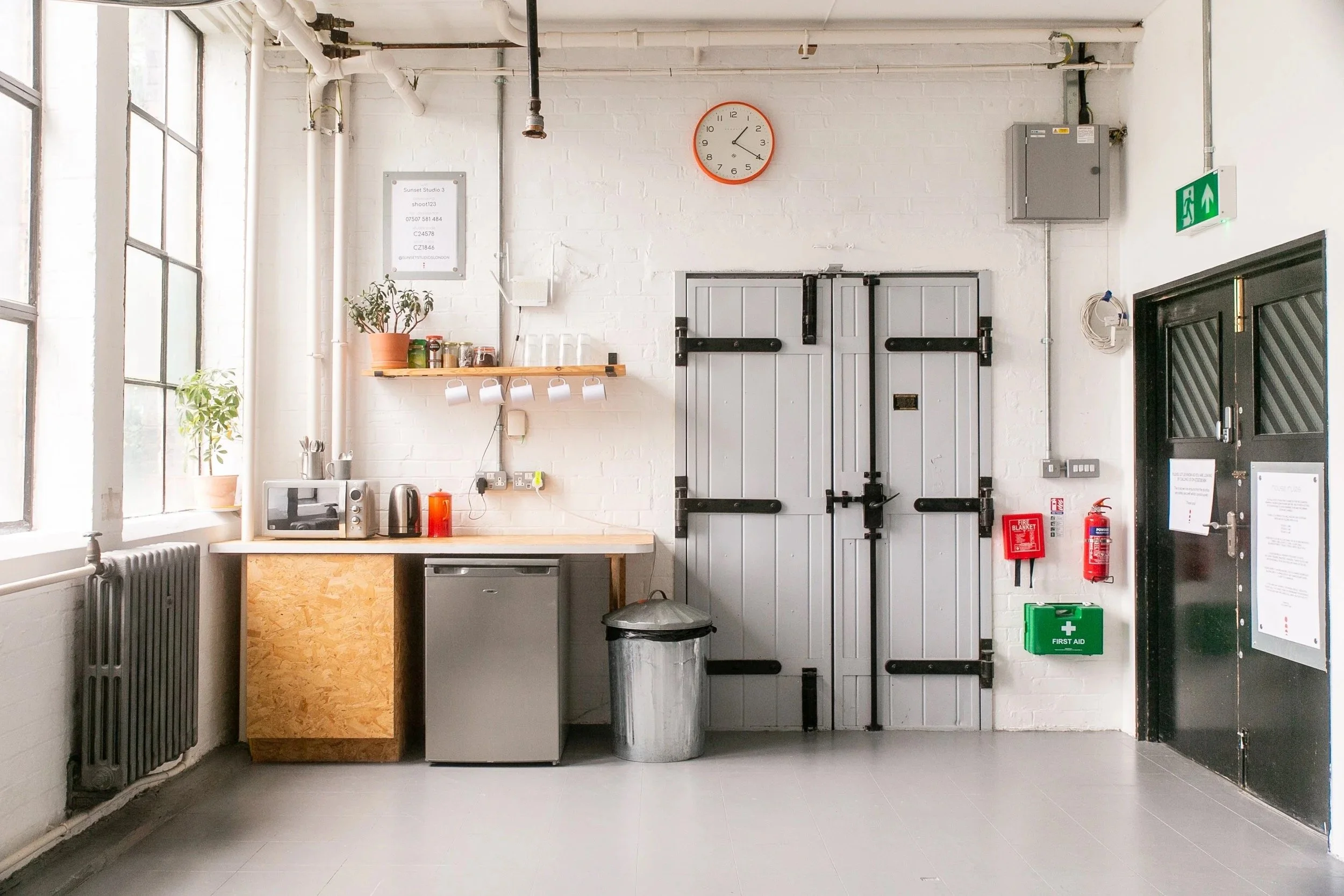 Inside a minimalist kitchen with white brick walls, a large window on the left, a round orange wall clock above, a small countertop with a microwave, kettle, and toaster, a potted plant, an open shelf with jars and mugs, a gray door in the middle, an