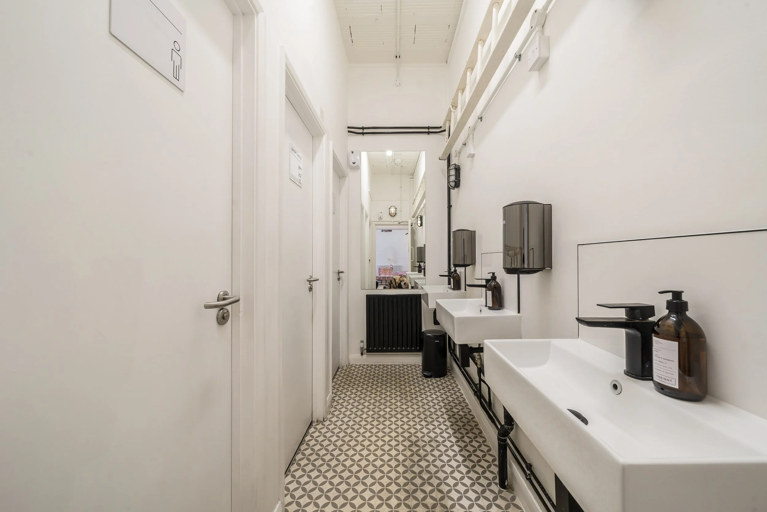 Public restroom with three white sinks, black soap dispensers, paper towel boxes, and black trash cans. White walls and patterned tile floor. Doors on left side, large mirror at the end of the room reflecting a seating area.